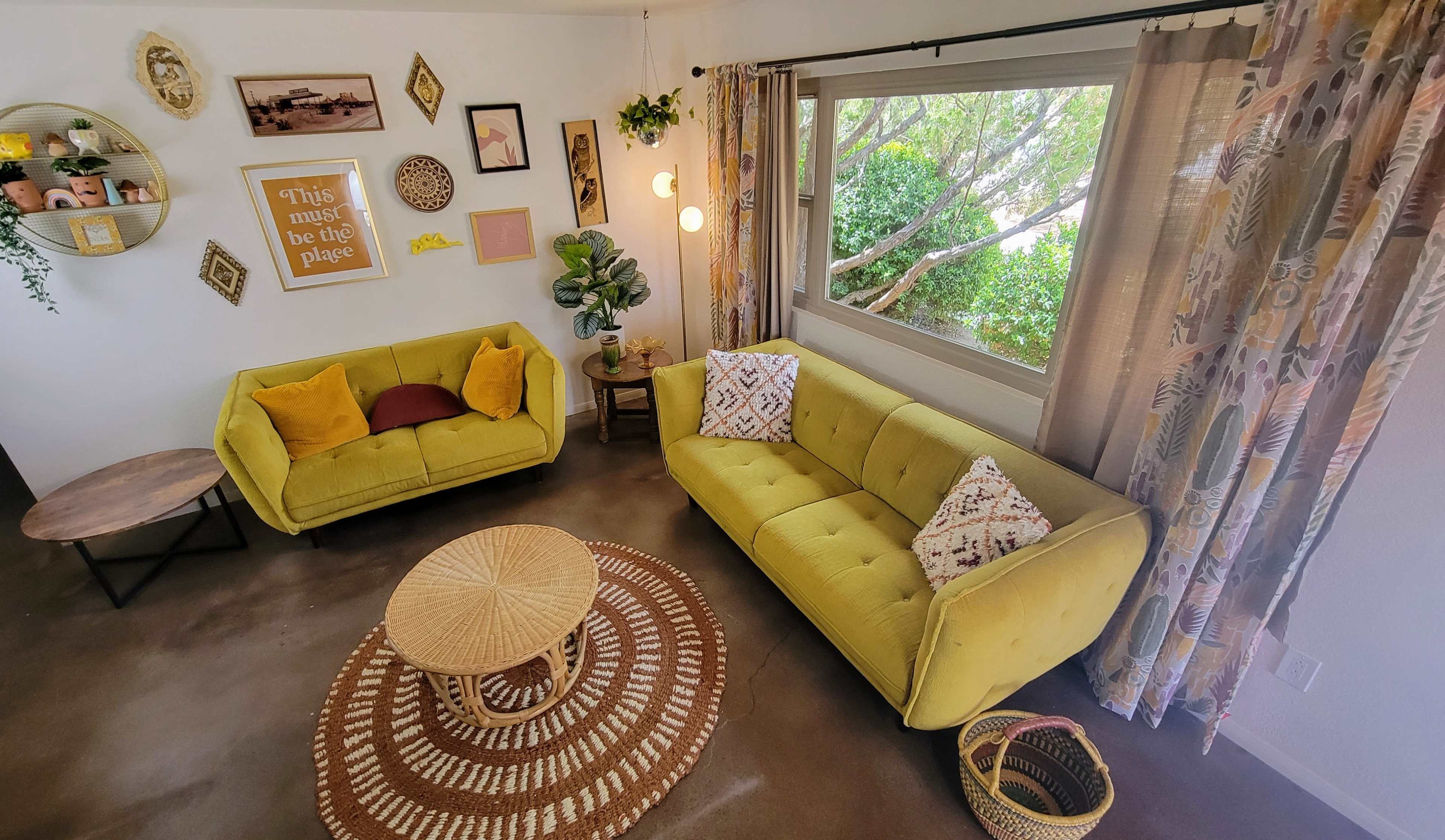 The image shows a cozy living room with two yellow sofas, a round coffee table, and a large window overlooking greenery.