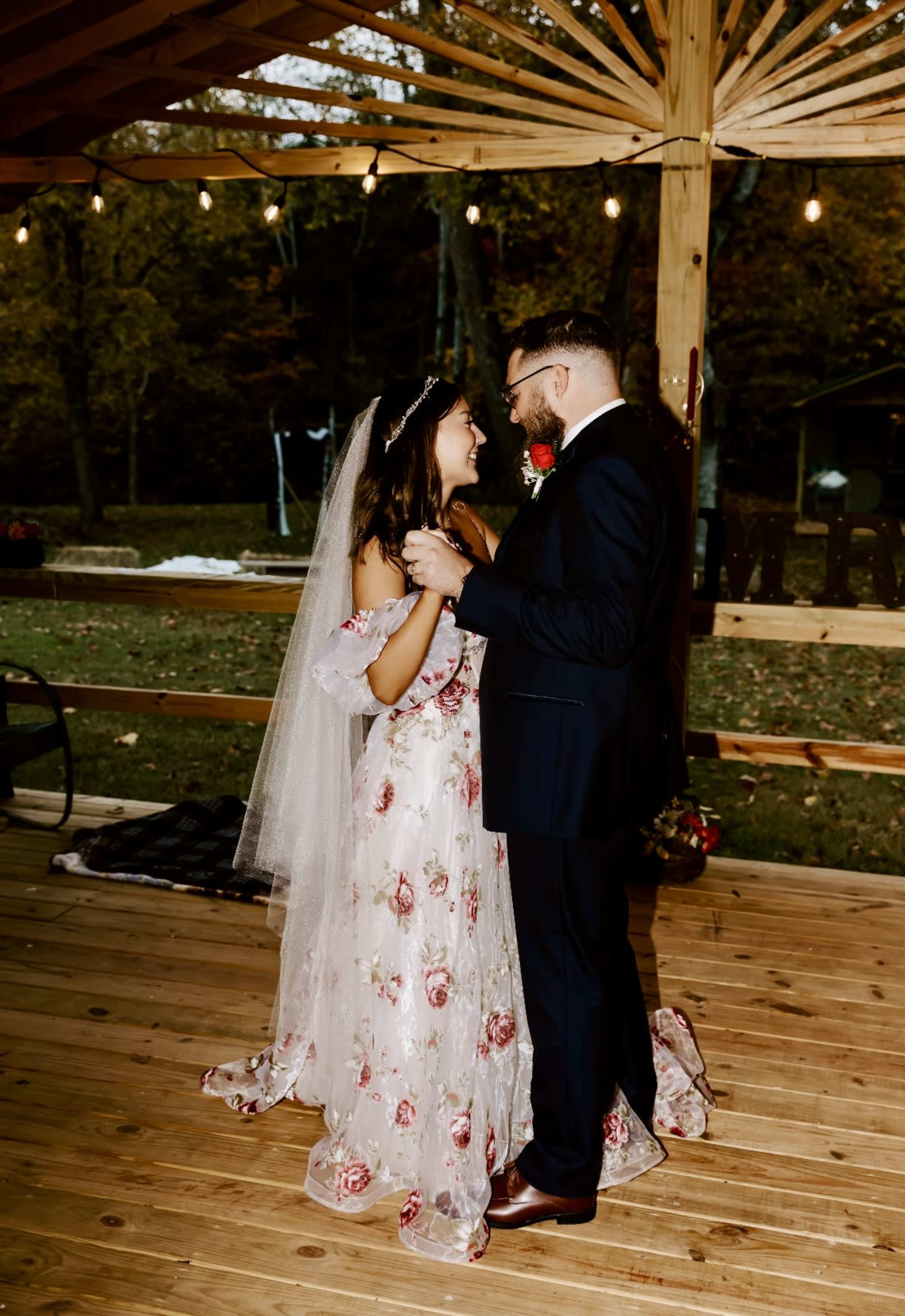A couple dances together under a wooden canopy adorned with string lights, surrounded by autumn foliage.