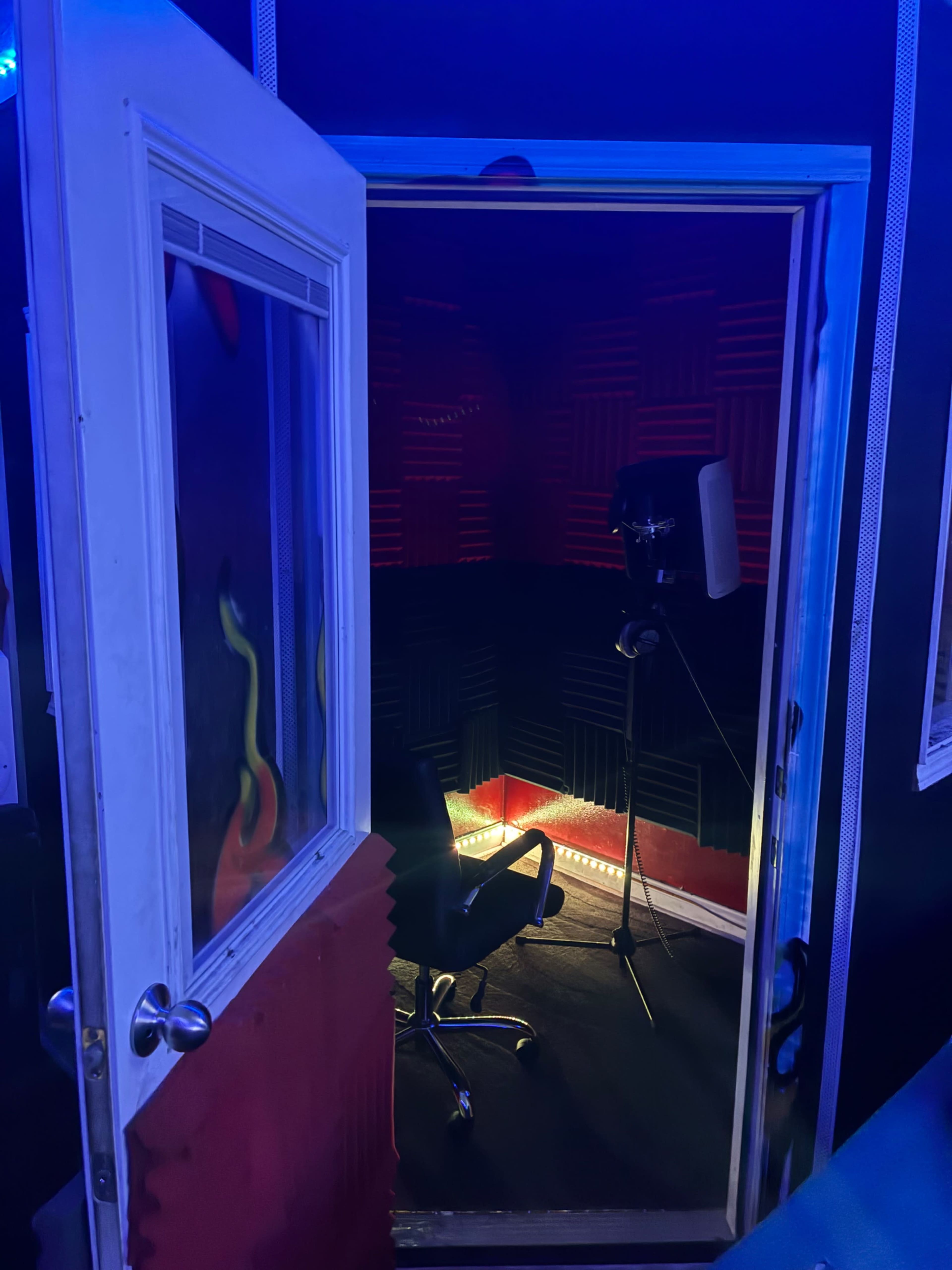 A soundproof recording studio is visible through an open door, featuring foam paneling, a microphone on a stand, and a chair in the center.