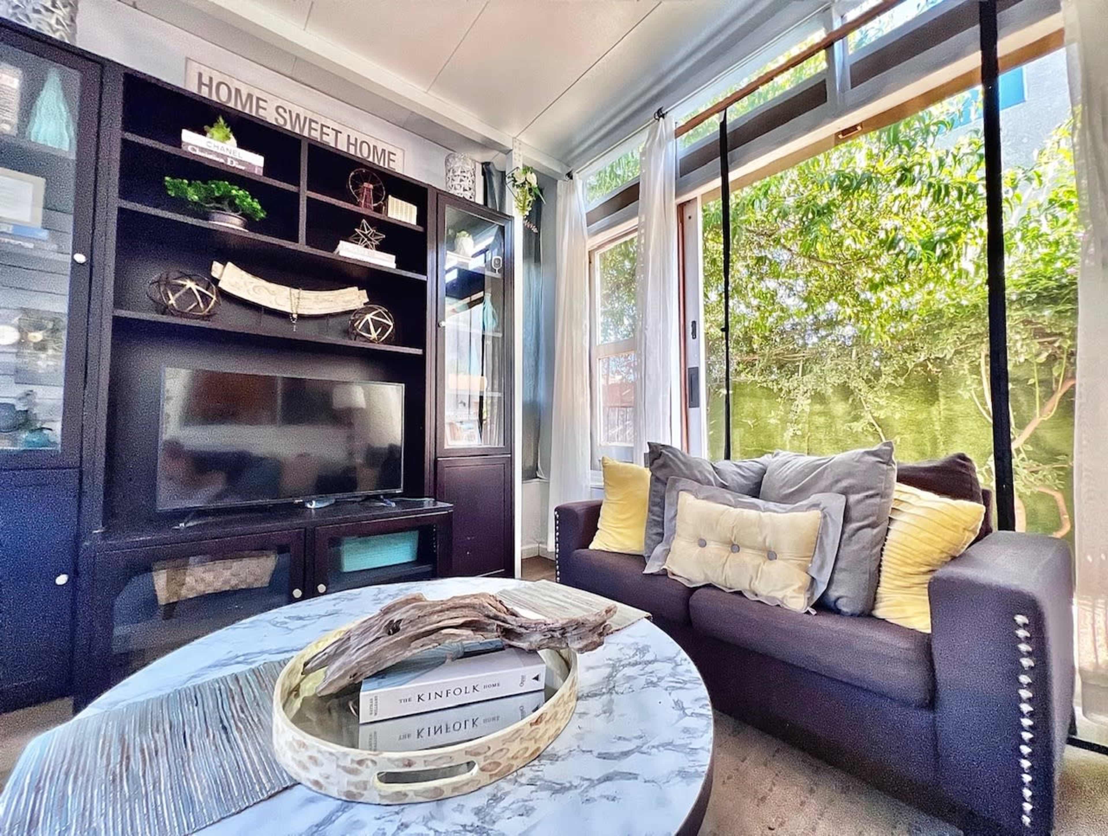 A cozy living room features a dark sofa with decorative pillows, a marble coffee table with books, and large windows showcasing greenery outside.