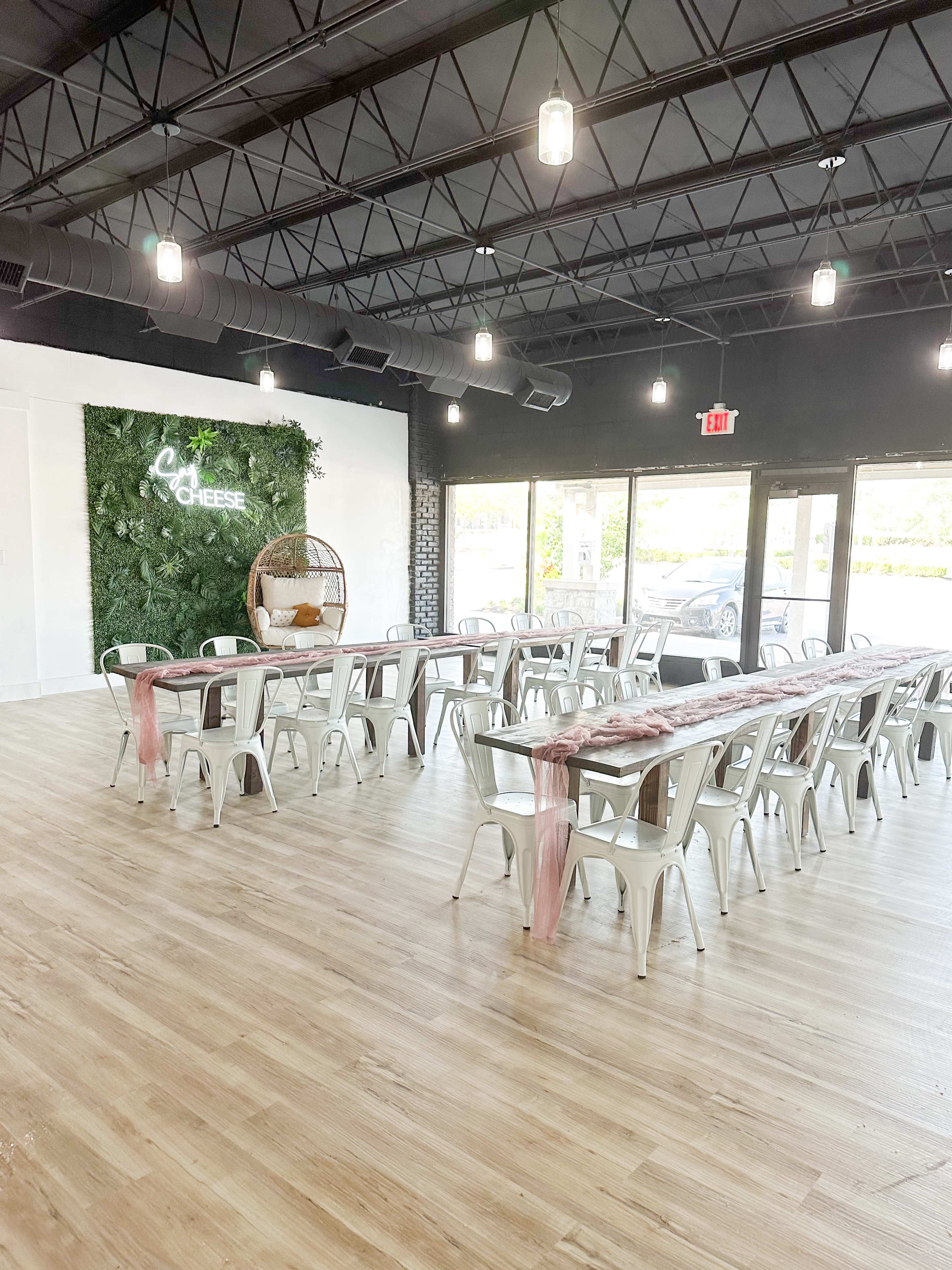 A spacious event venue with long tables arranged in rows, decorated with pink runners, and a green wall featuring the words "Cheese."