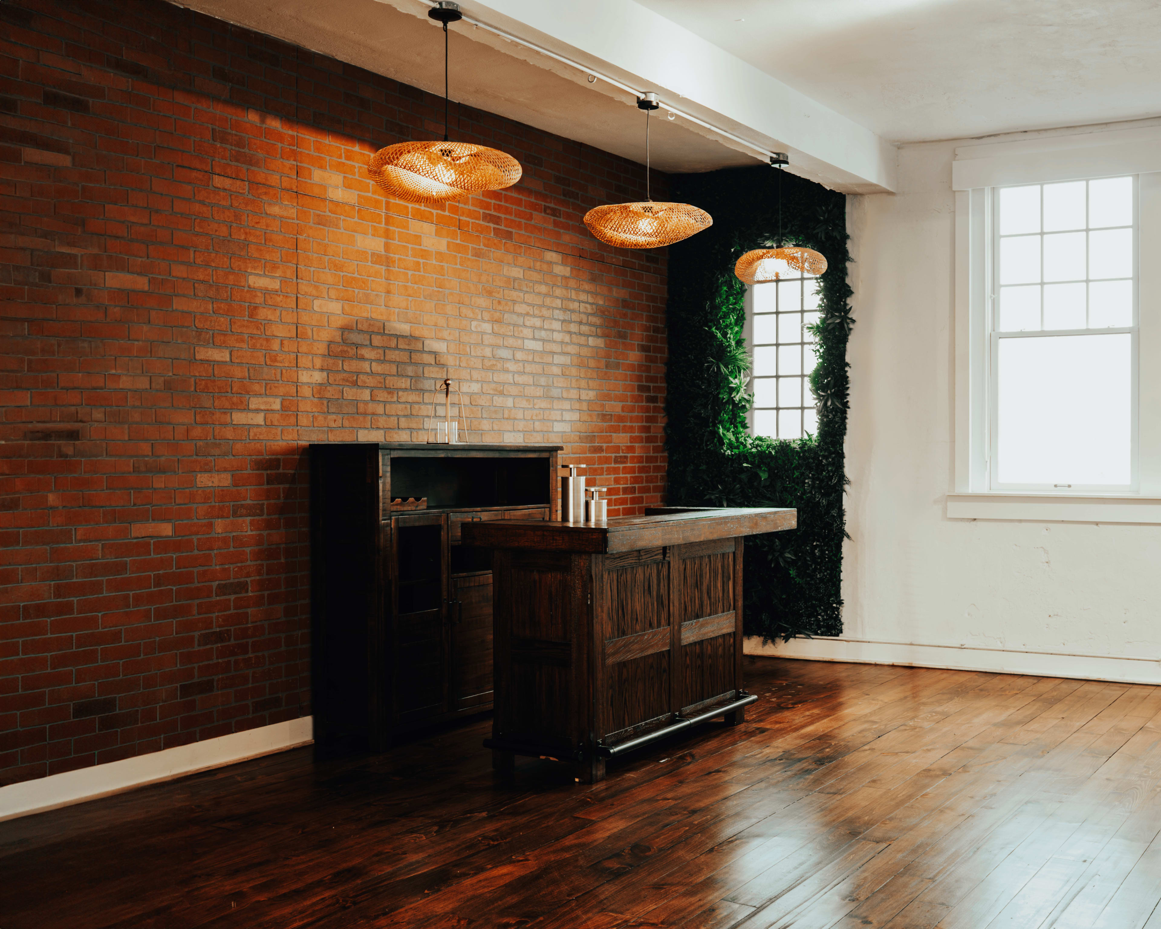 A modern, sparsely furnished room with a brick wall, wooden flooring, a bar counter, and hanging pendant lights.