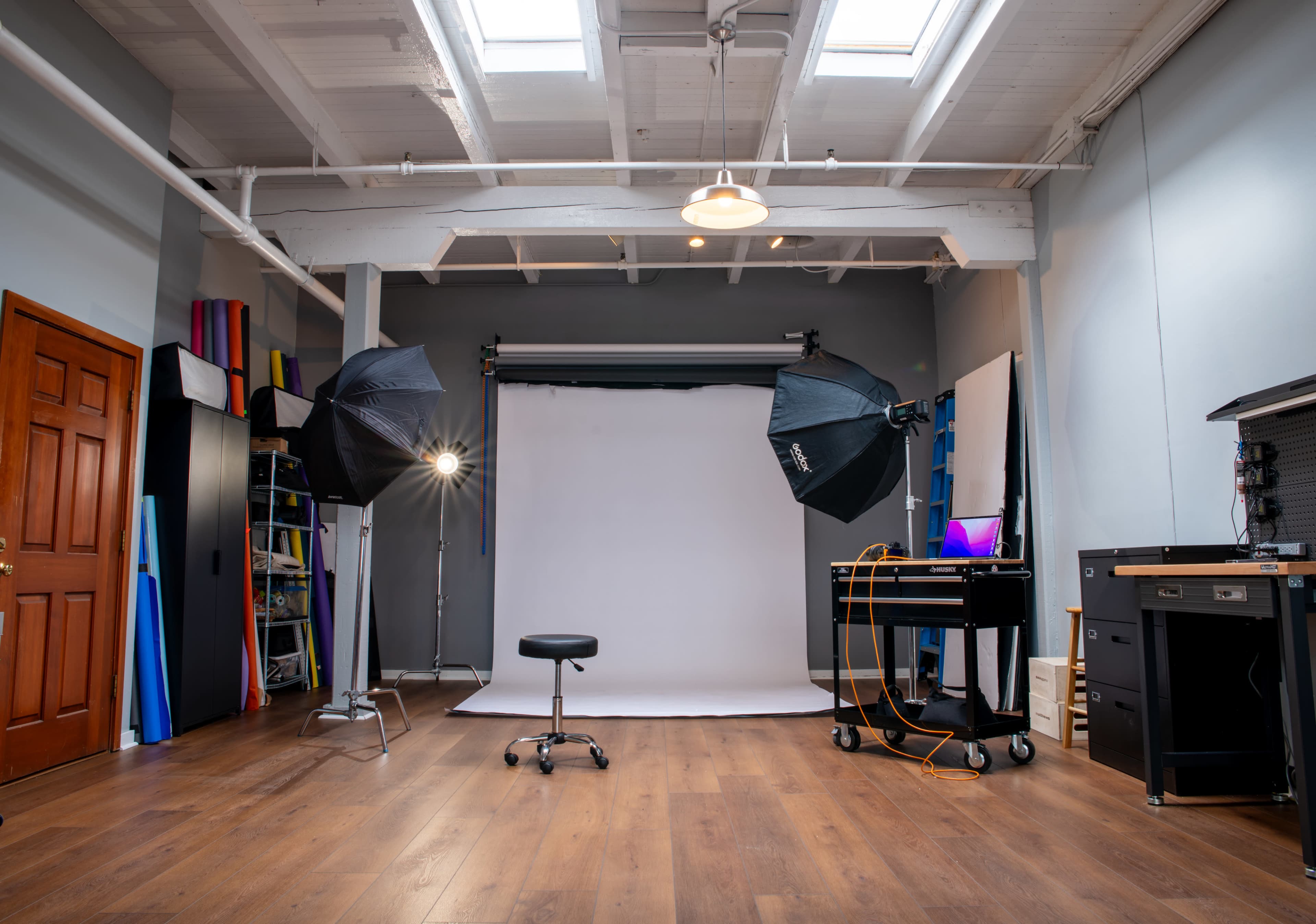 The image shows a photography studio with a white backdrop, two large studio lights, and a stool in the center, surrounded by various equipment and colorful backdrops on the walls.