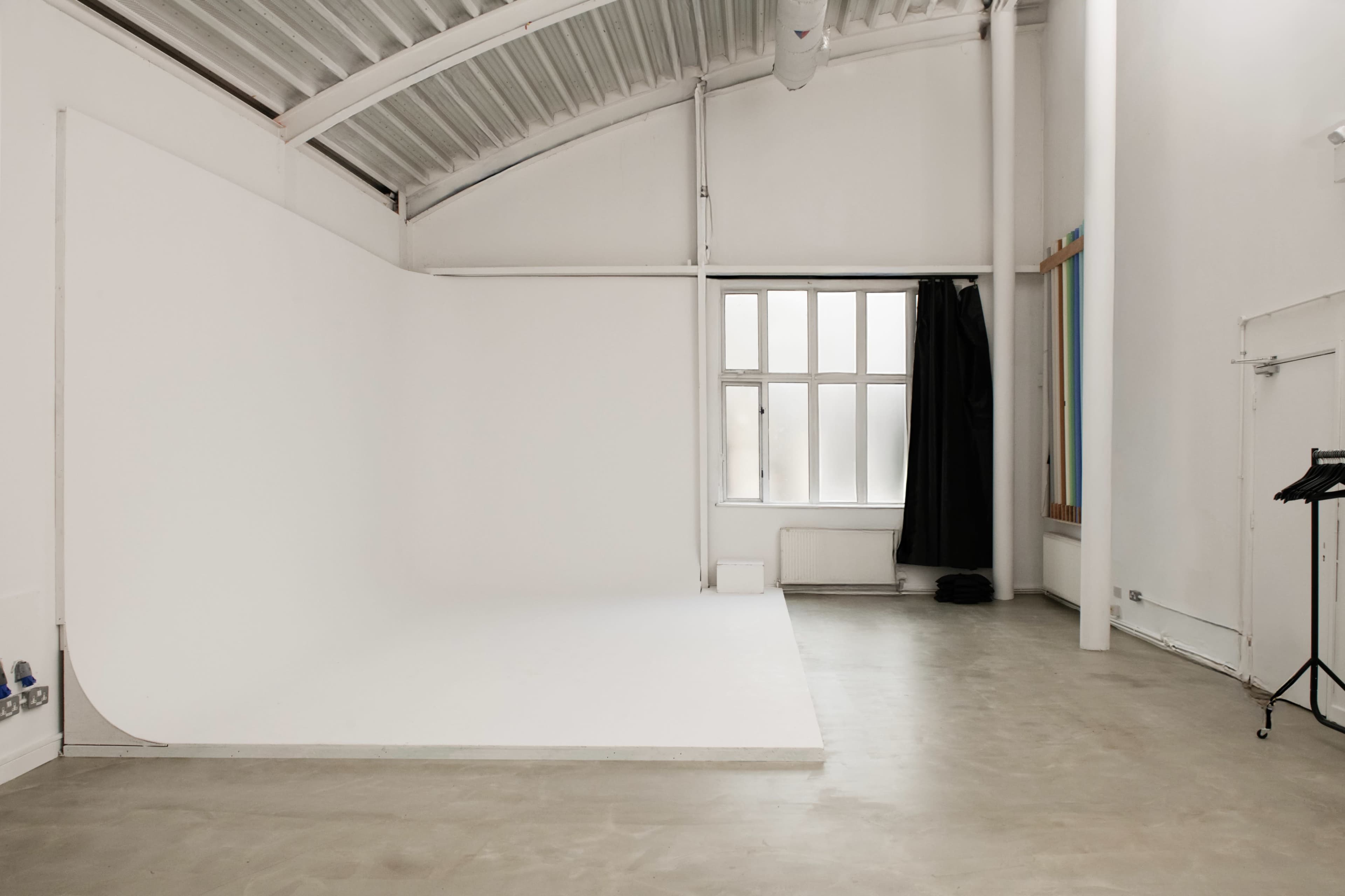 The image shows a spacious photography studio with a white seamless backdrop, large windows, and a concrete floor.