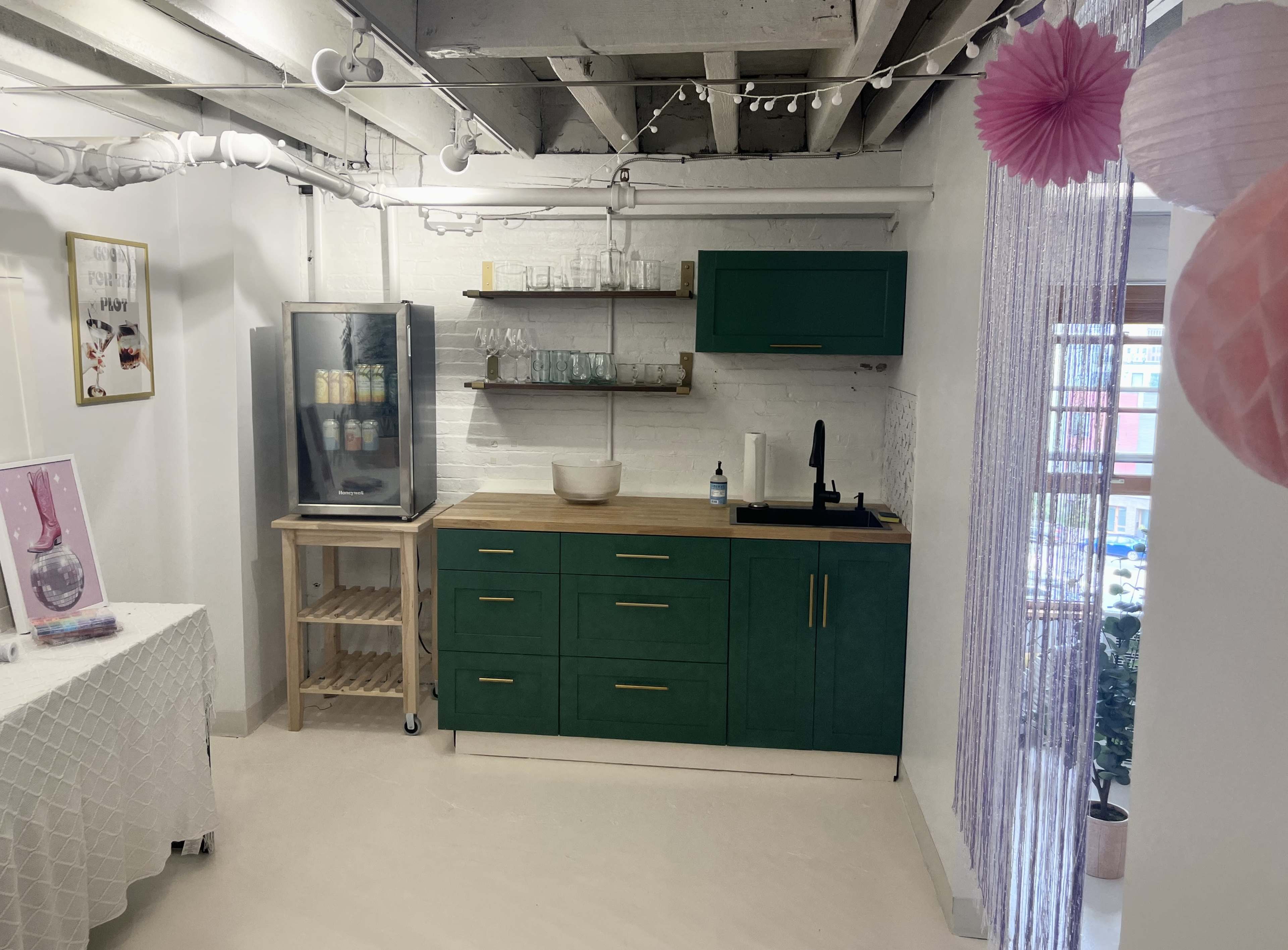 A modern kitchen area with green cabinetry, a wooden countertop, and a glass refrigerator, complemented by decorative elements like pink paper lanterns and a white tablecloth.