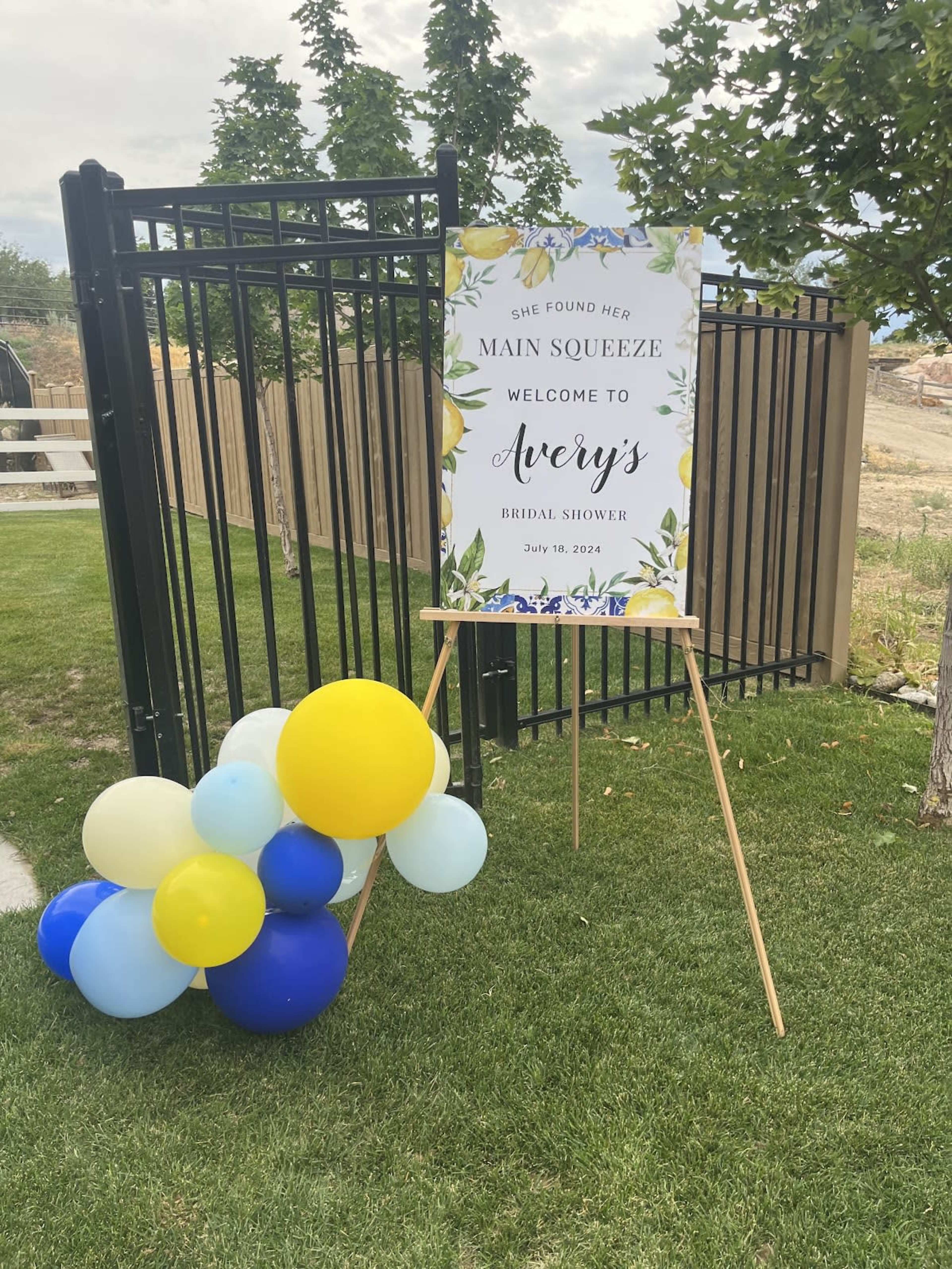 A sign announcing a bridal shower for someone named Avery is displayed on an easel beside a cluster of blue, yellow, and light blue balloons.