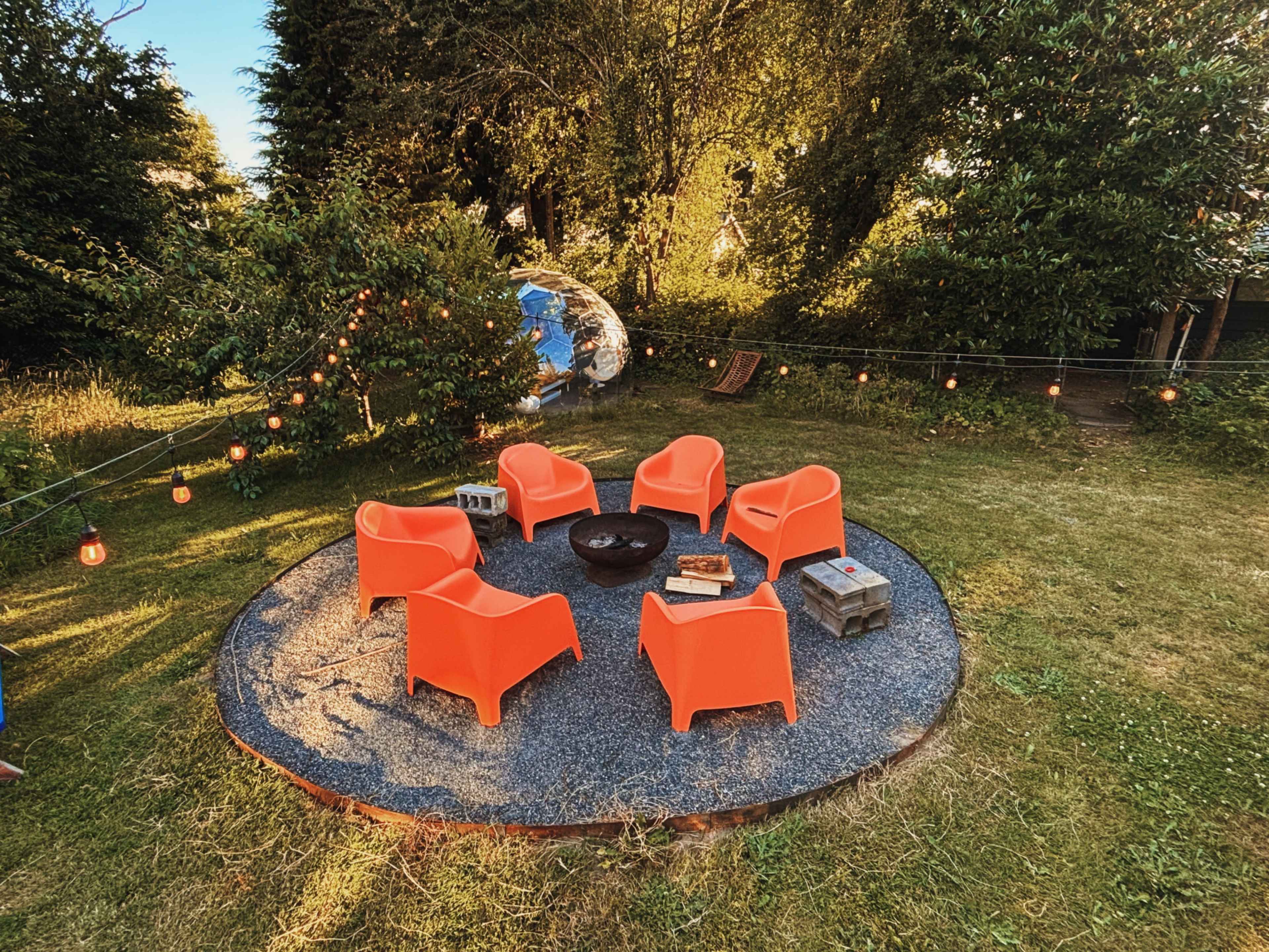 A circular arrangement of five orange chairs surrounds a fire pit on a gravel patio in a grassy yard illuminated by string lights.