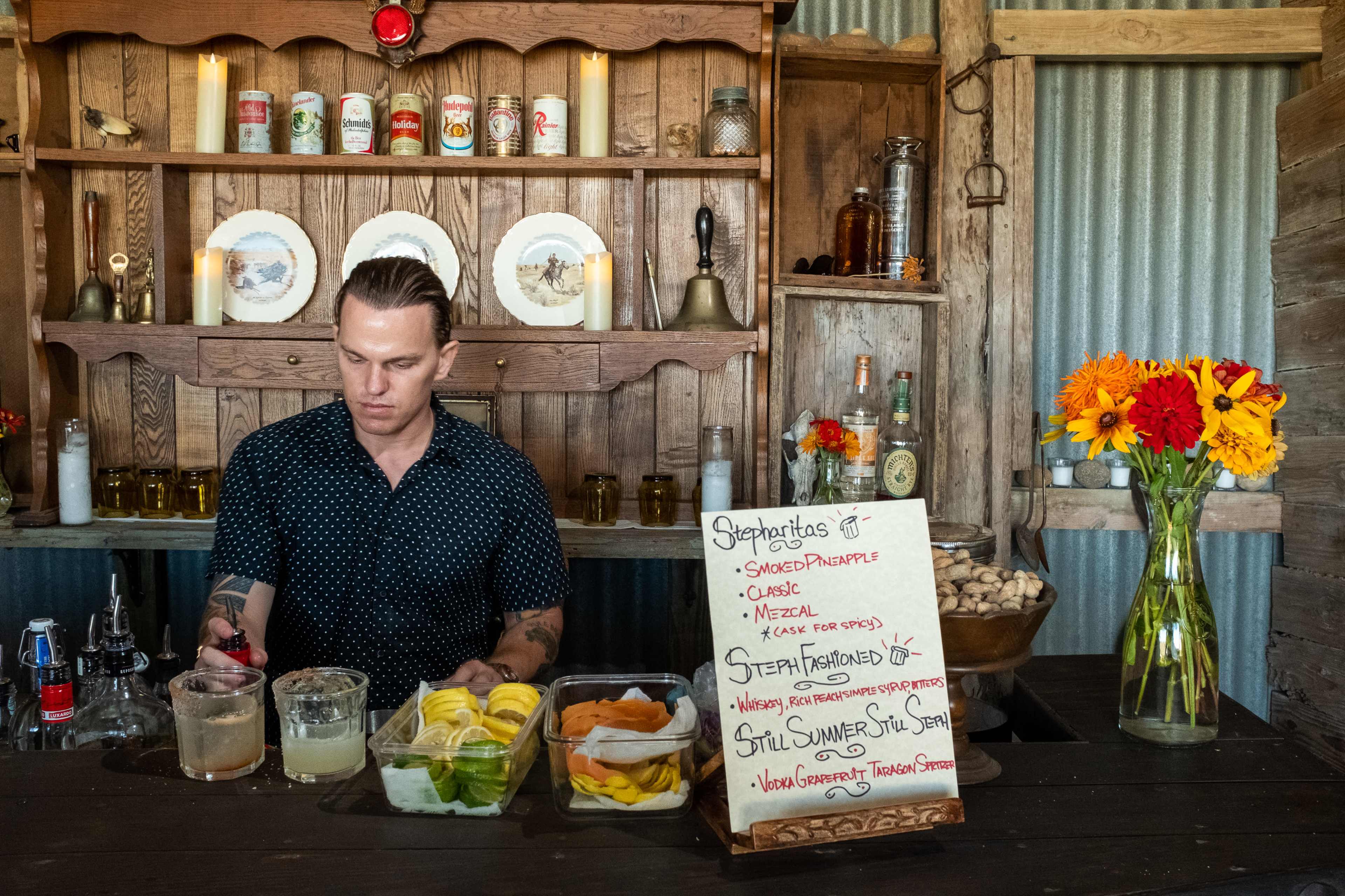 A bartender prepares drinks at a rustic bar with a variety of fruits and a menu displayed on a wooden board.