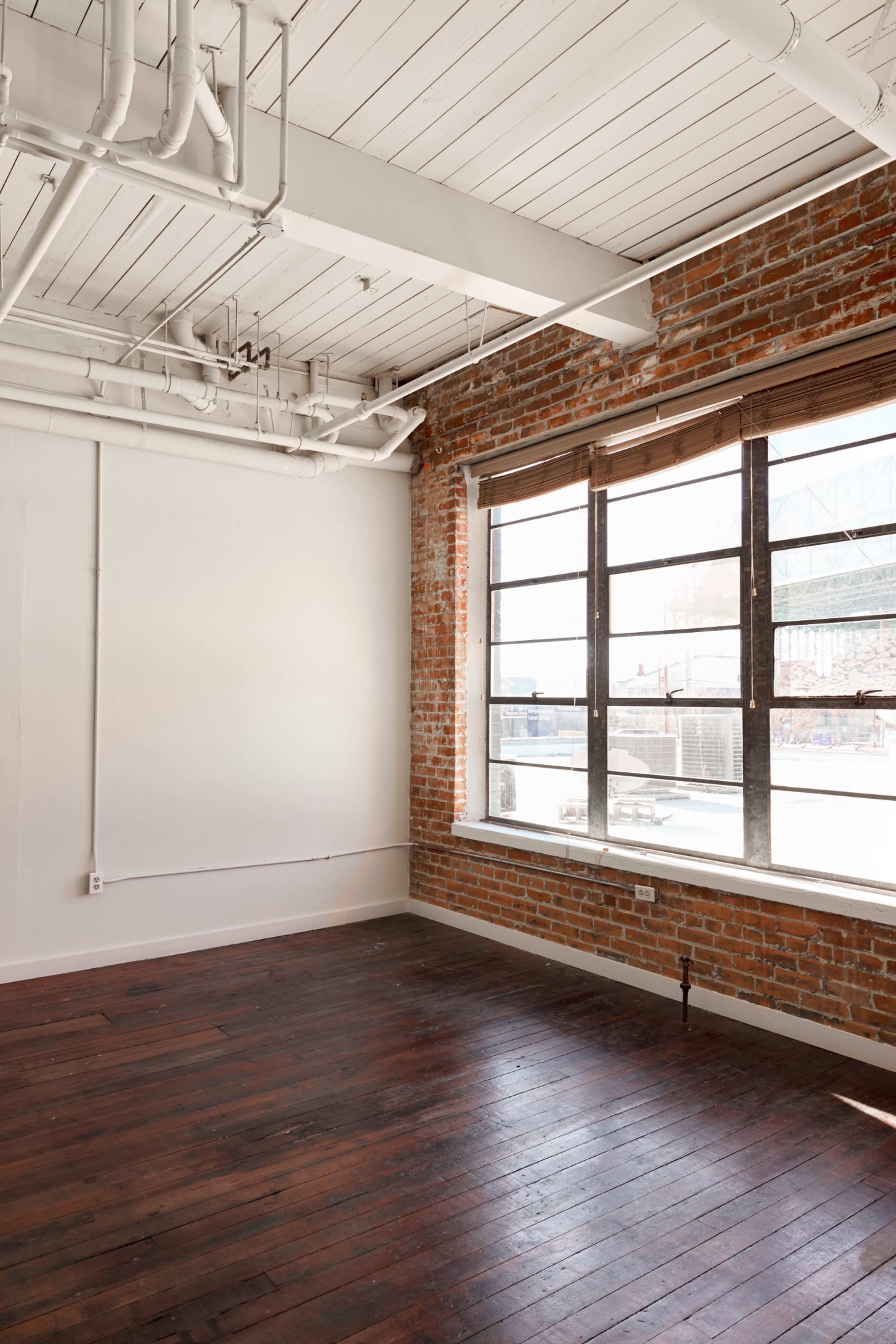 An empty room with exposed brick walls, large windows, and wooden flooring.