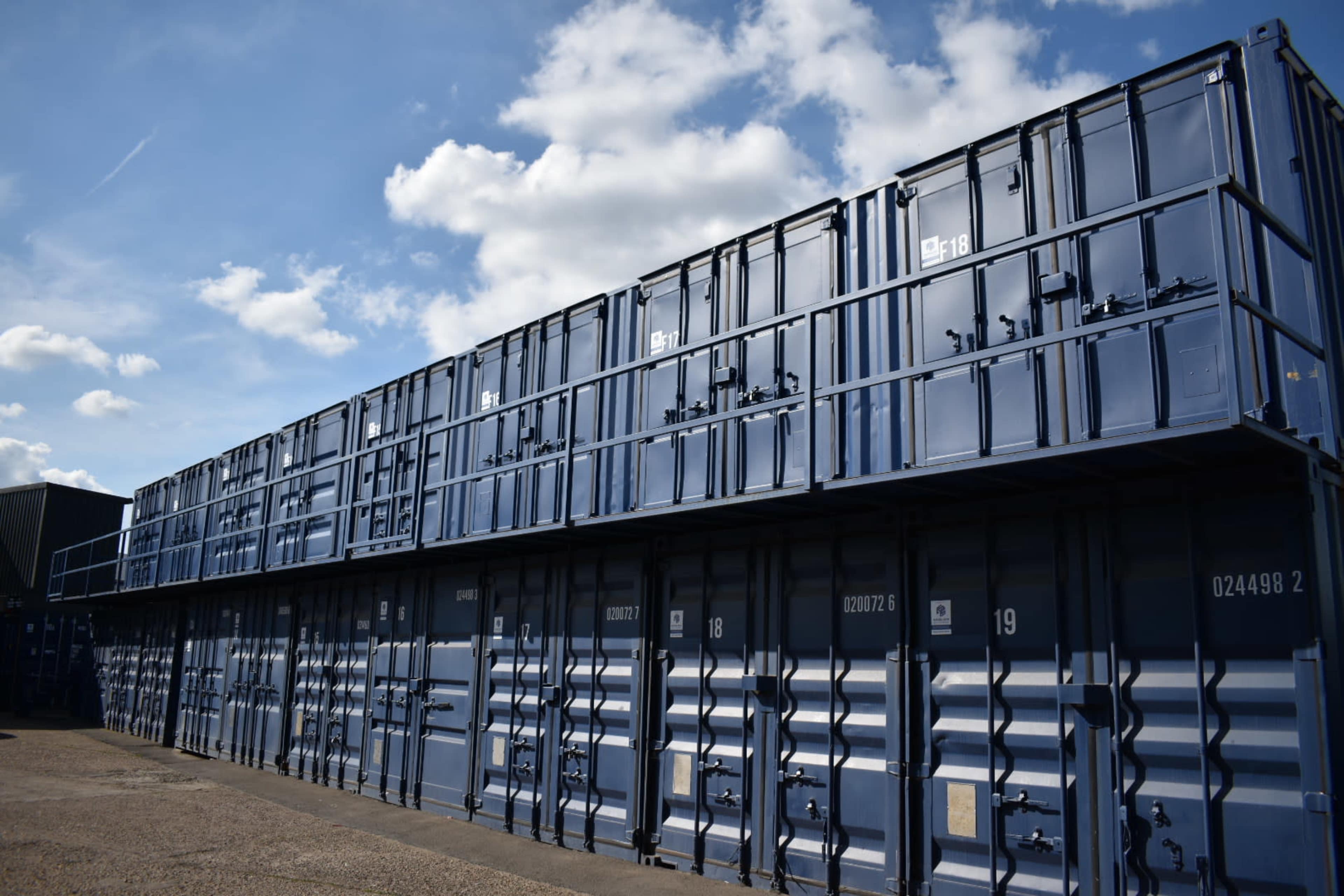 The image shows a row of stacked blue shipping containers against a partly cloudy sky.