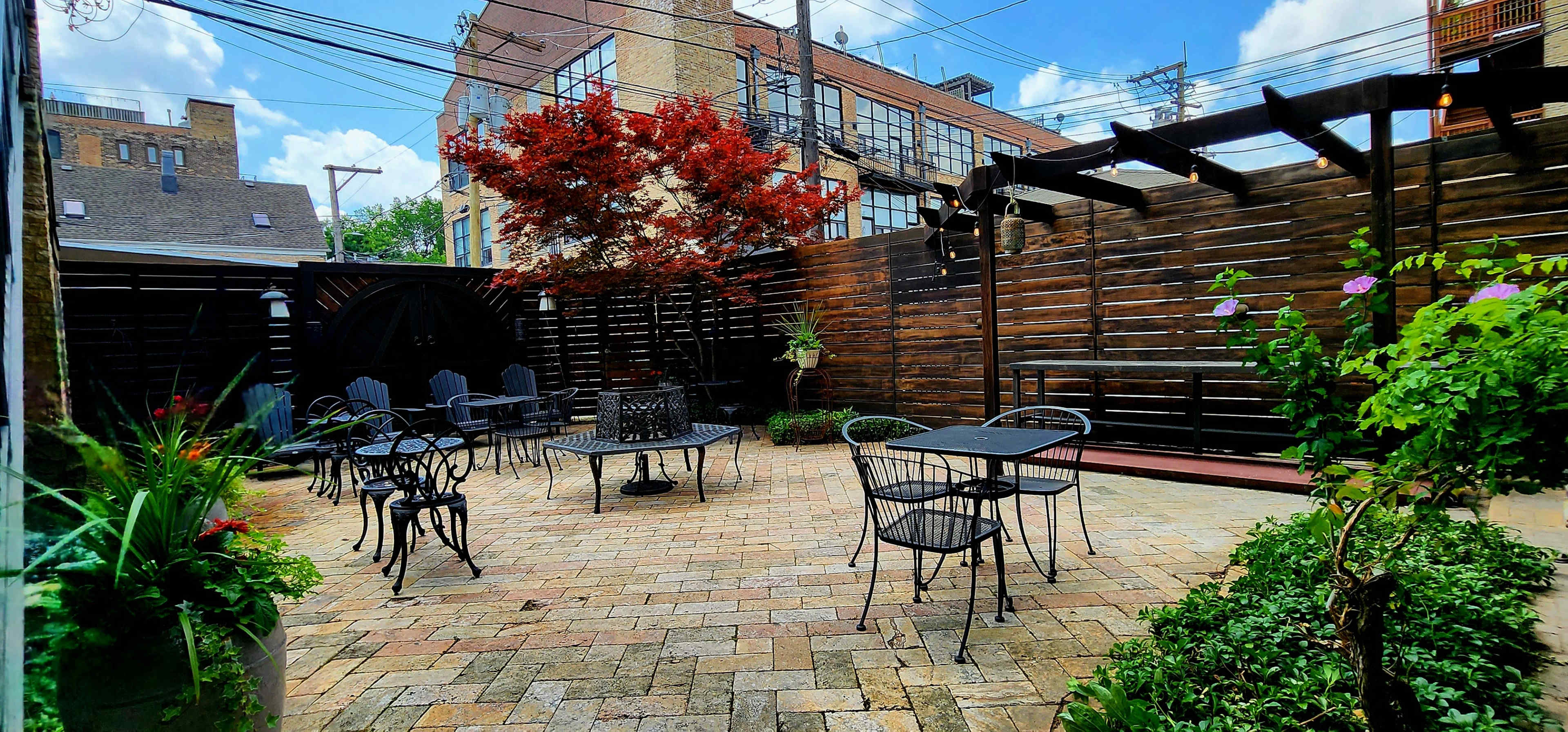 A brick-paved patio features multiple metal tables and chairs surrounded by a wooden fence and potted plants.