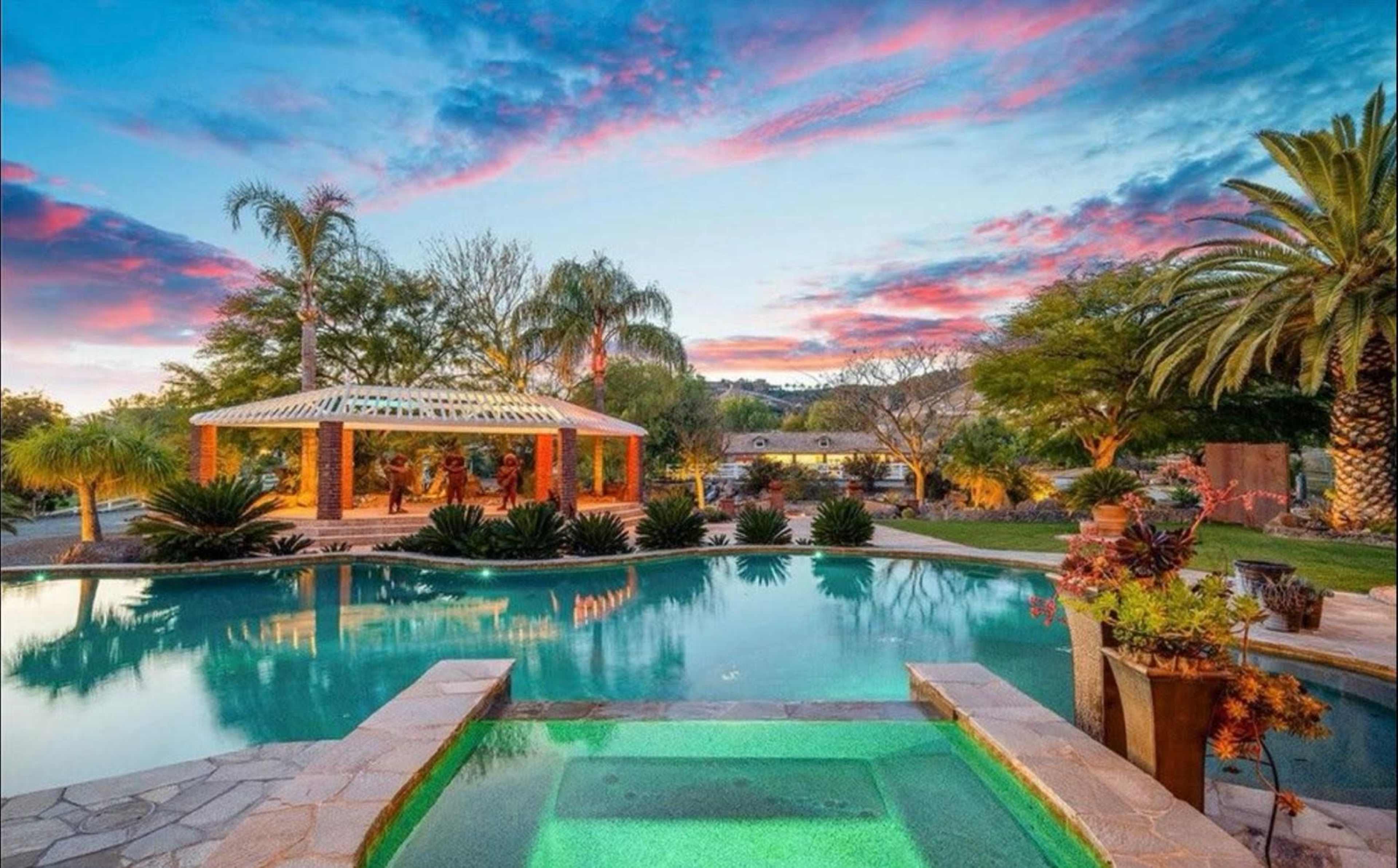 A backyard oasis features a swimming pool and spa surrounded by lush landscaping and a gazebo, all under a colorful sunset sky.