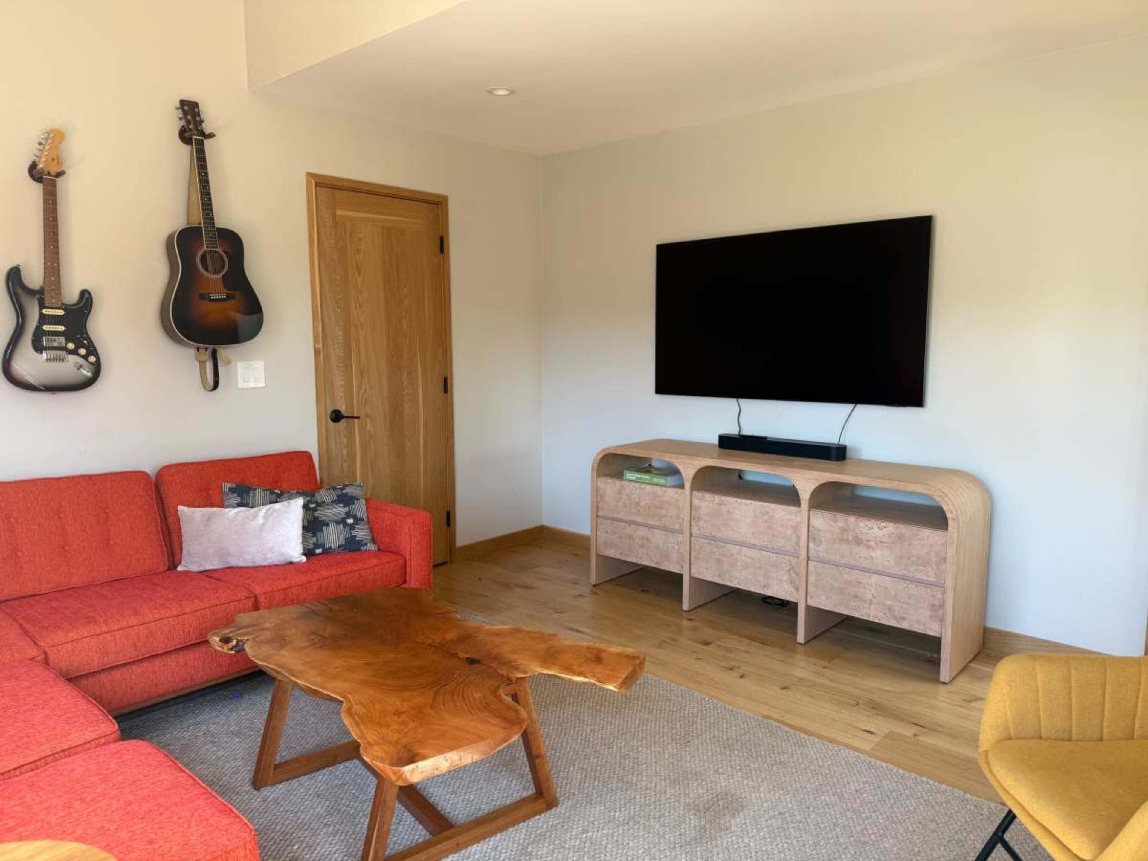 The room features a red couch, a wooden coffee table, a TV mounted on the wall, and two guitars hanging beside a wooden door.