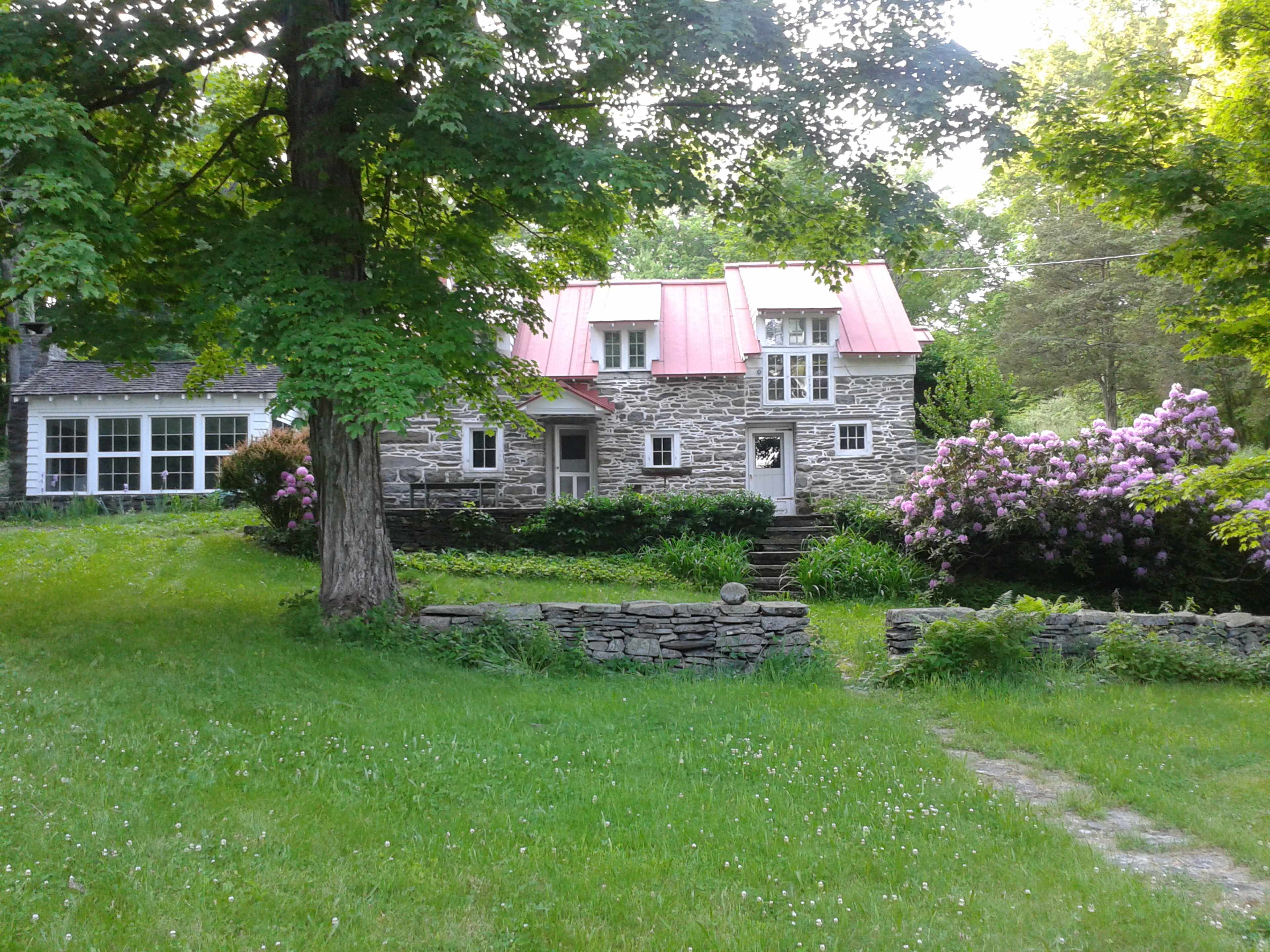 A stone house with a red metal roof is surrounded by green grass and blooming shrubs.