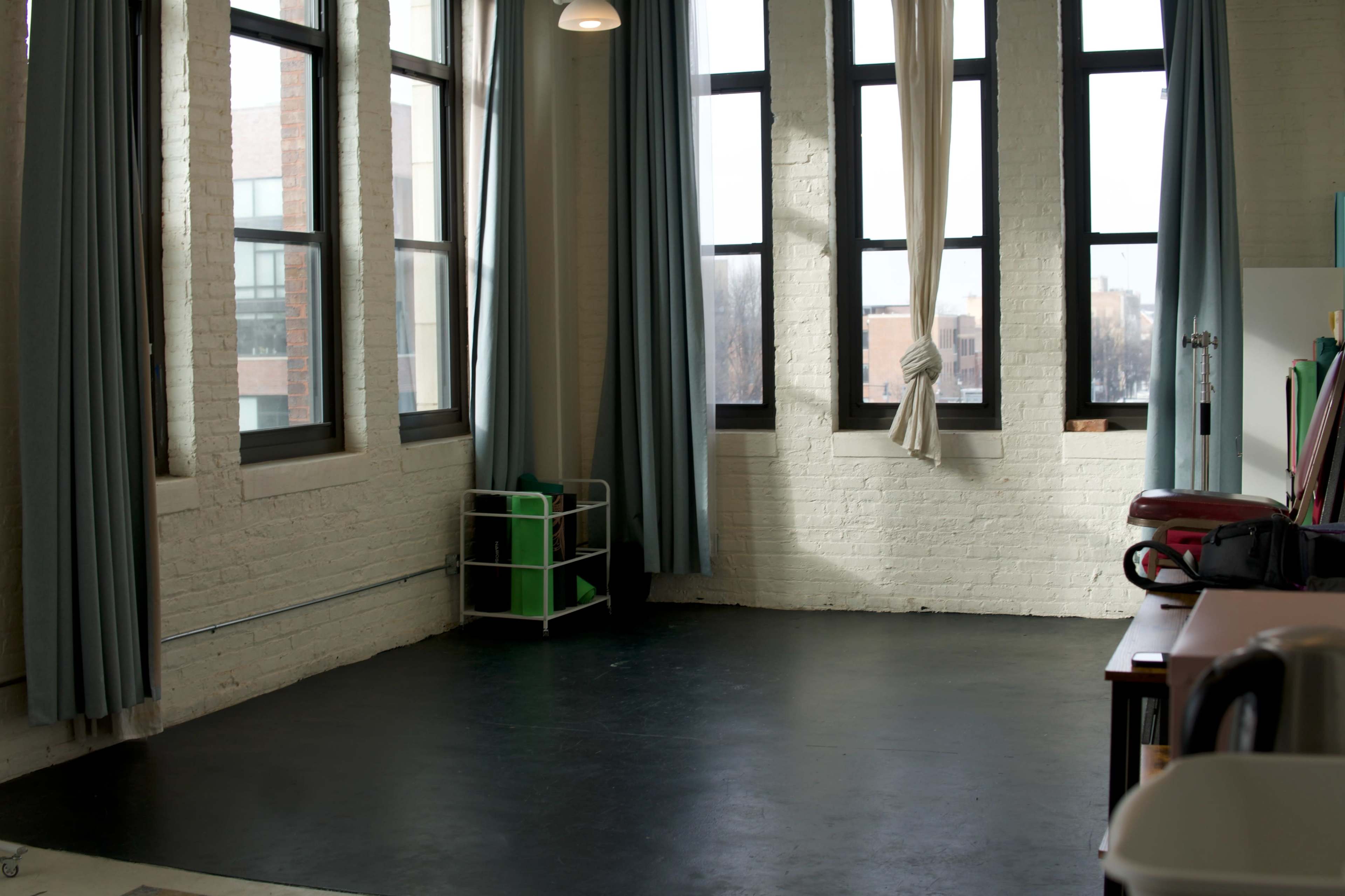 The image shows a spacious room with large windows, exposed brick walls, and a black floor, featuring a green storage cart in one corner.