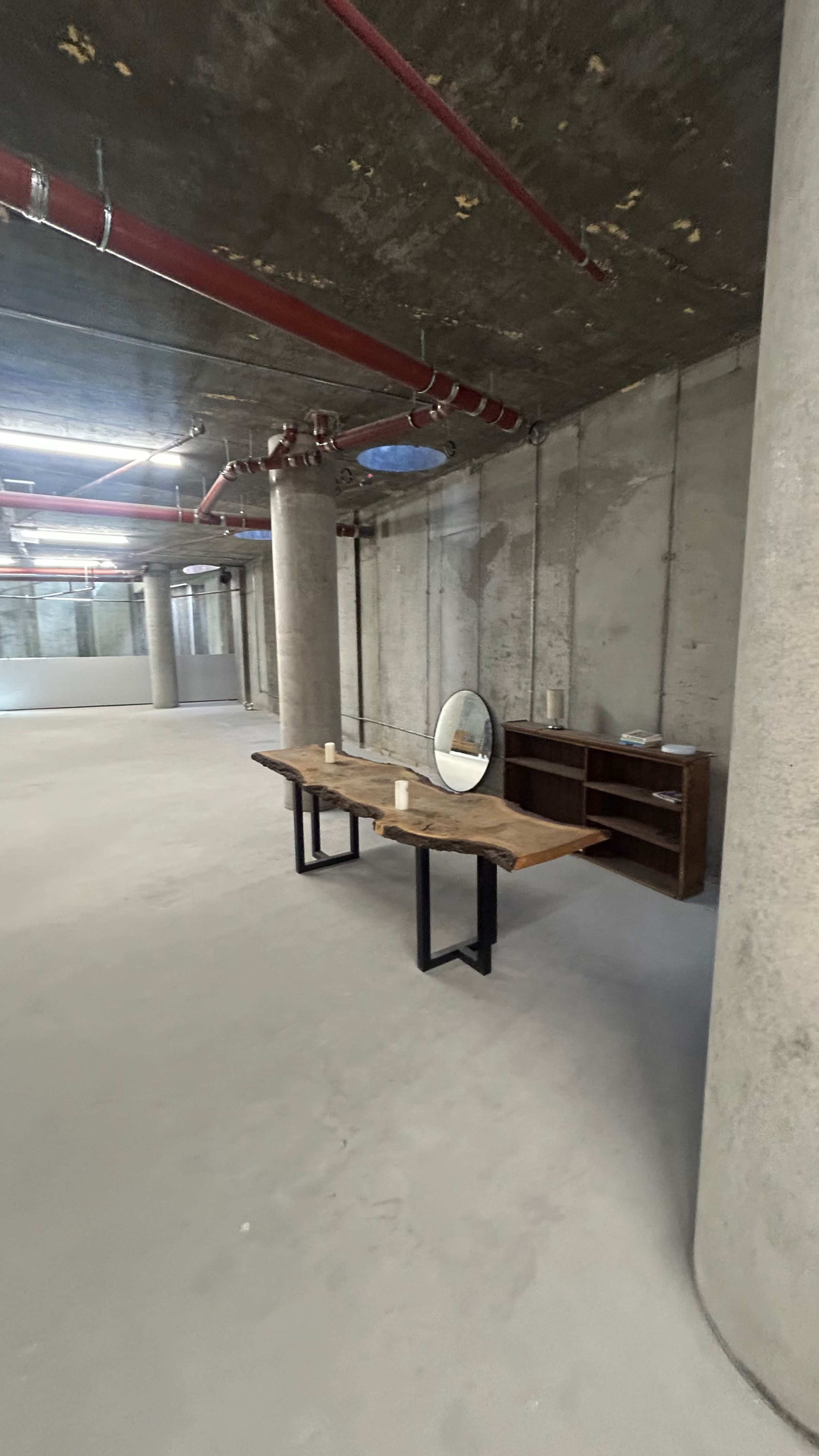 The image shows a large, empty room with concrete walls and pillars, featuring a wooden table with a mirror and a wooden shelf against one wall.