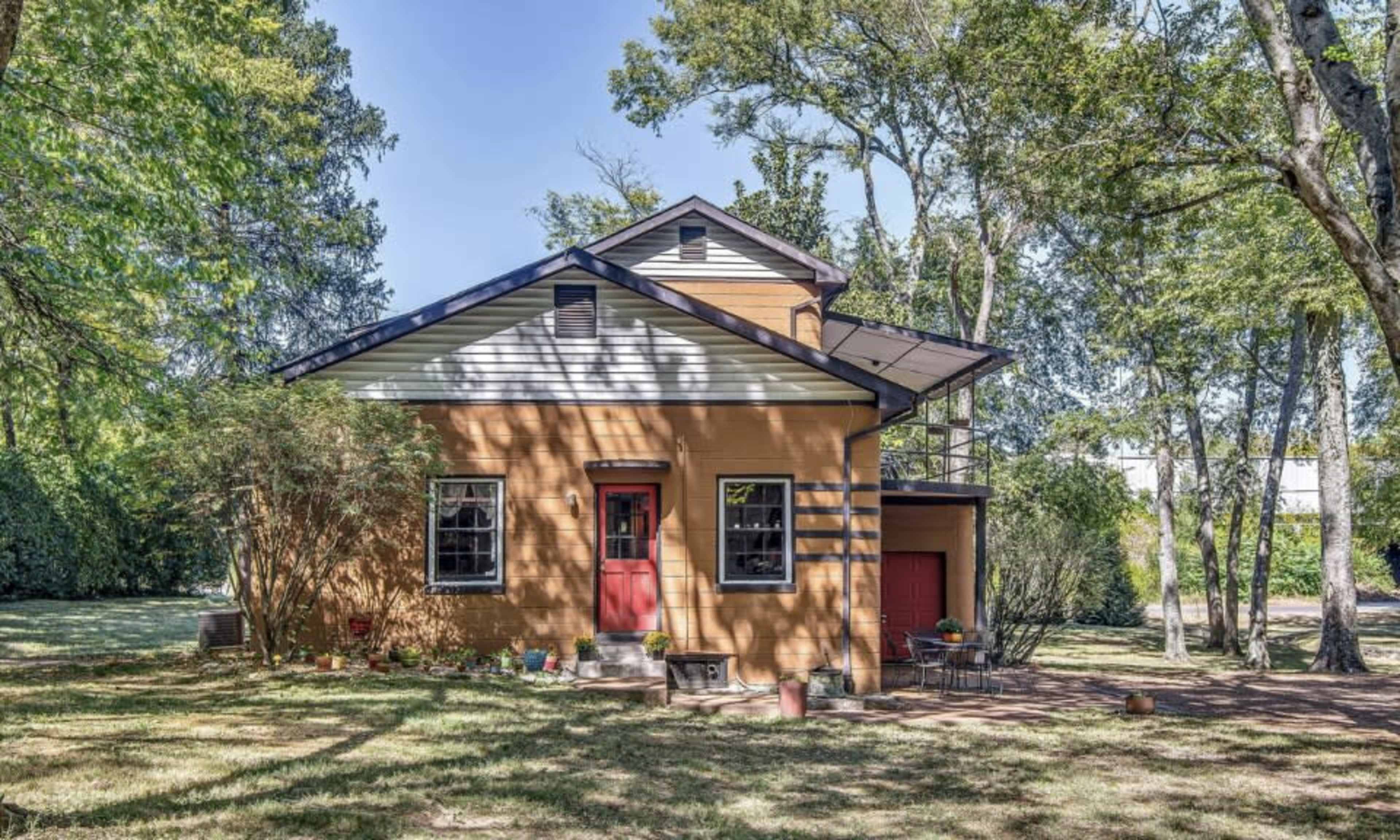 The image shows a two-story house with a brown exterior, a slanted roof, and red doors, surrounded by trees and a grassy area.