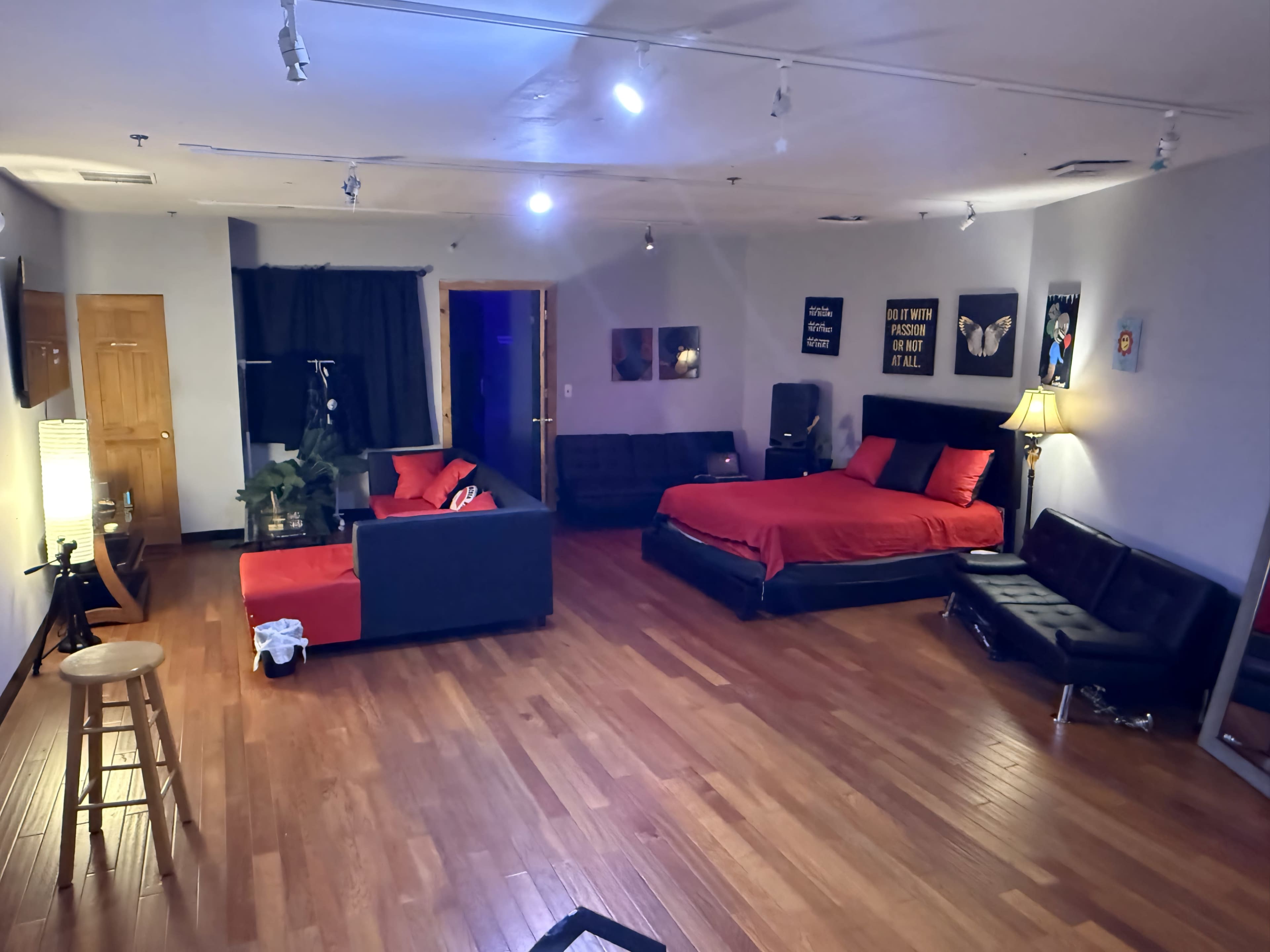 The image shows a spacious room with wooden floors, featuring a black-and-red color scheme, a bed, a sofa set, and various artworks on the walls.