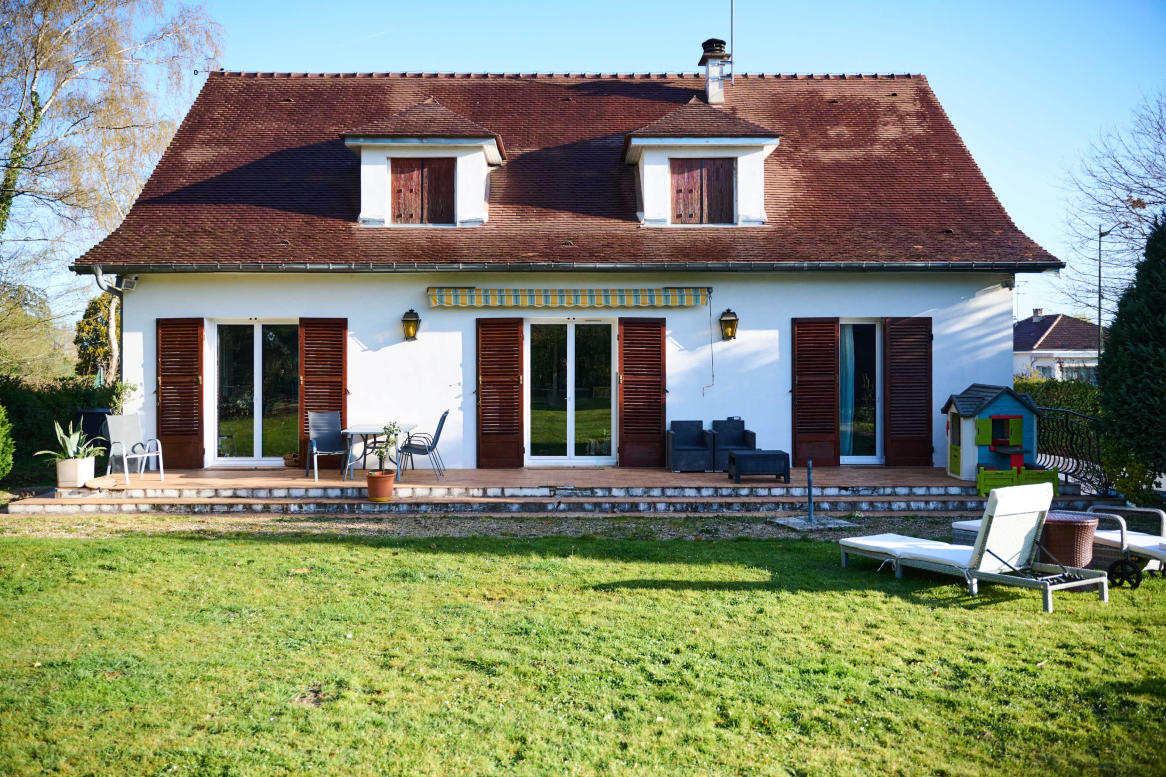 A two-story house with a gabled roof features multiple shuttered windows, a patio, and a well-maintained lawn.