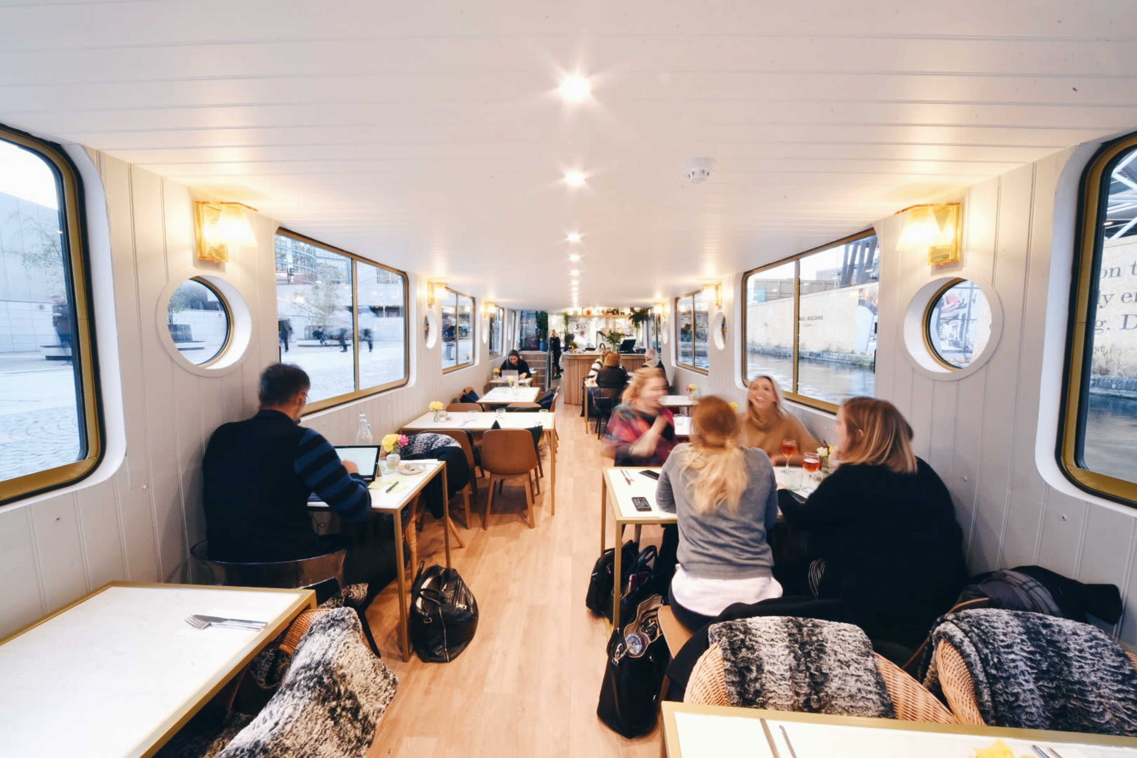 The interior of a boat restaurant features several groups of people dining at tables, with large round windows providing views of the water outside.