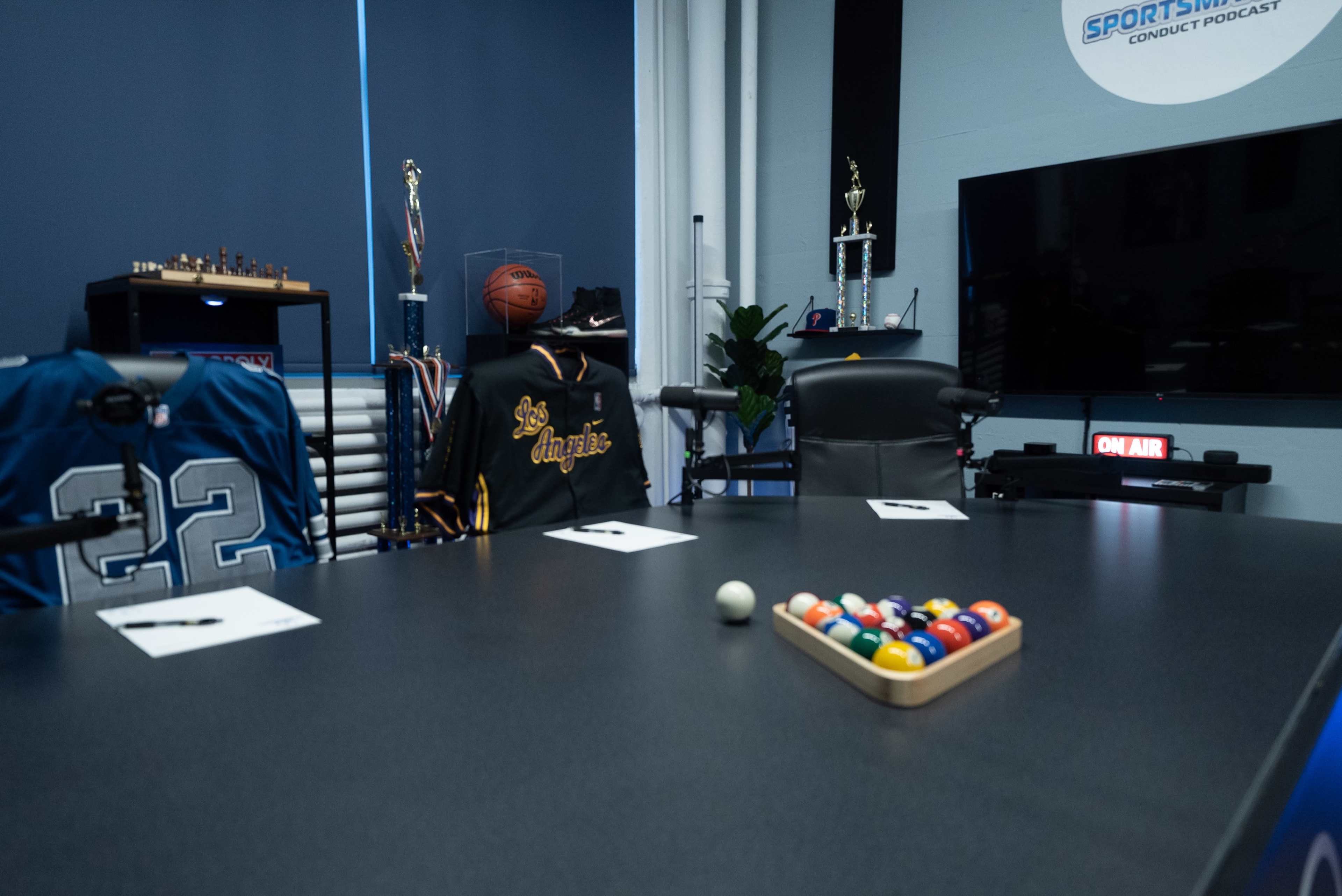 A conference room is set up with a pool table featuring a triangle of colored balls, jerseys on chairs, trophies displayed, and a visible "ON AIR" sign.