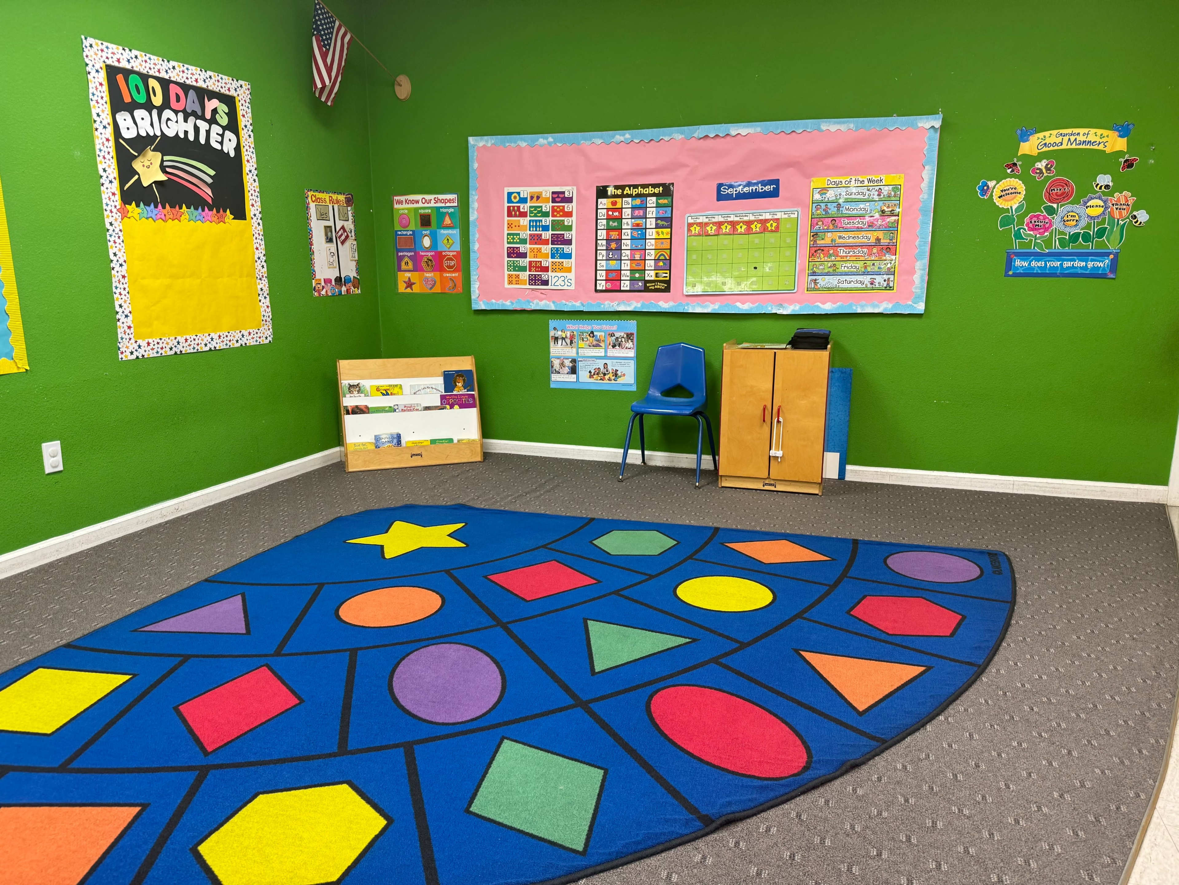 A classroom features a green wall, a large colorful rug with geometric shapes, a blue chair, and educational posters on the walls.