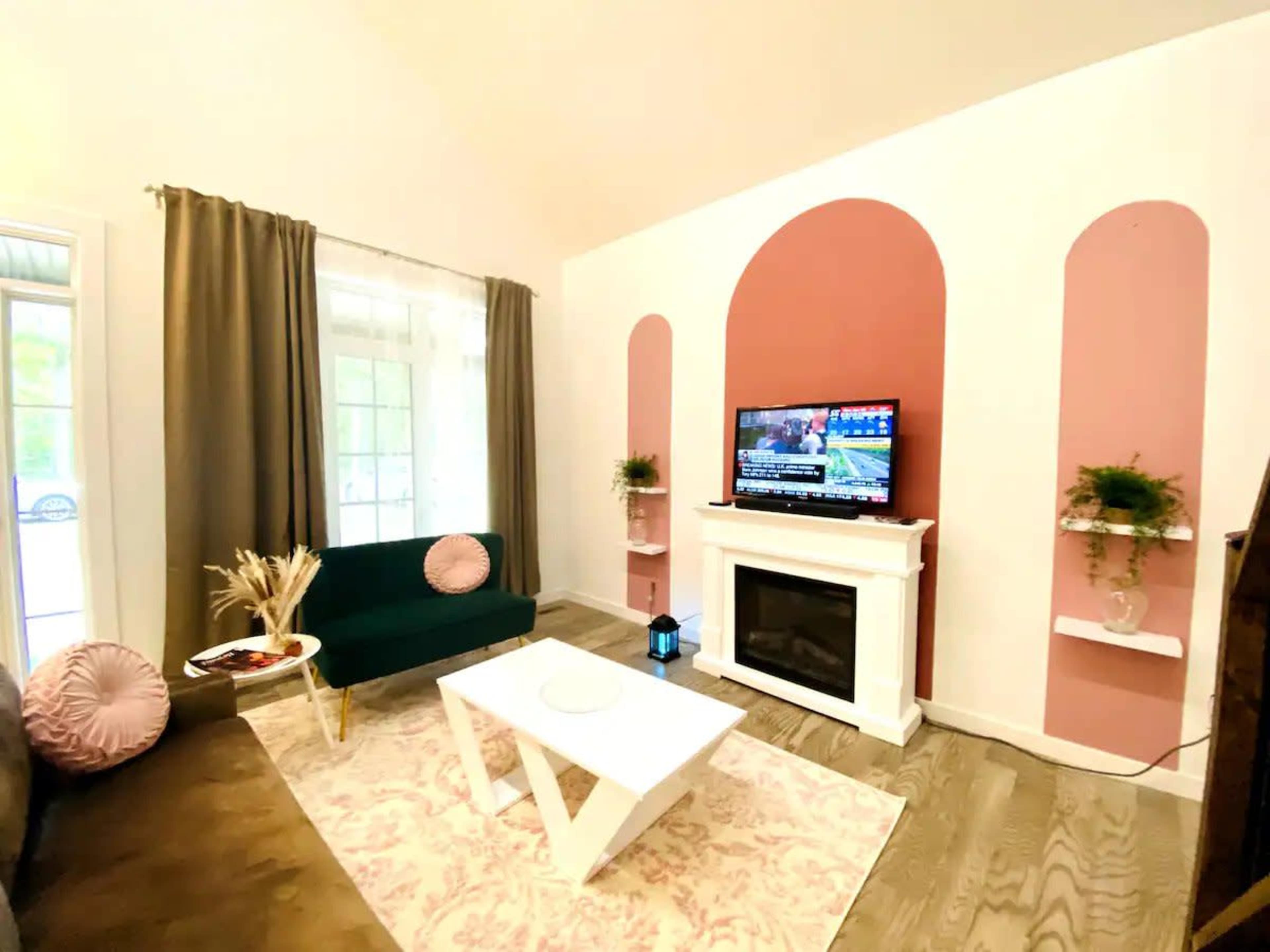 A living room with a green couch, a white coffee table, a television mounted on a pink-accented wall, and decorative plants on shelves.
