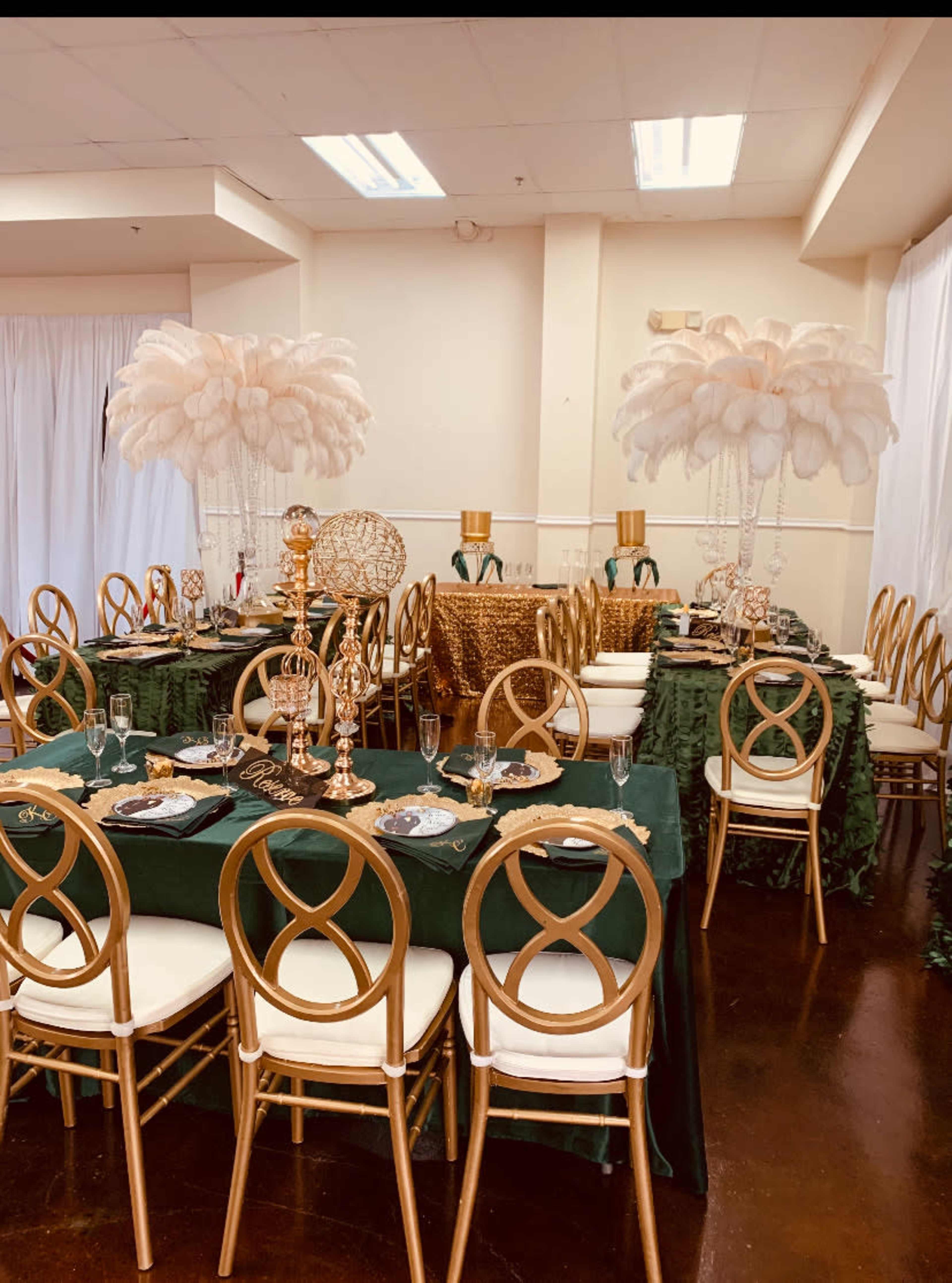 A banquet hall is set up with elegantly decorated tables featuring gold chairs, green tablecloths, and tall feather centerpieces.