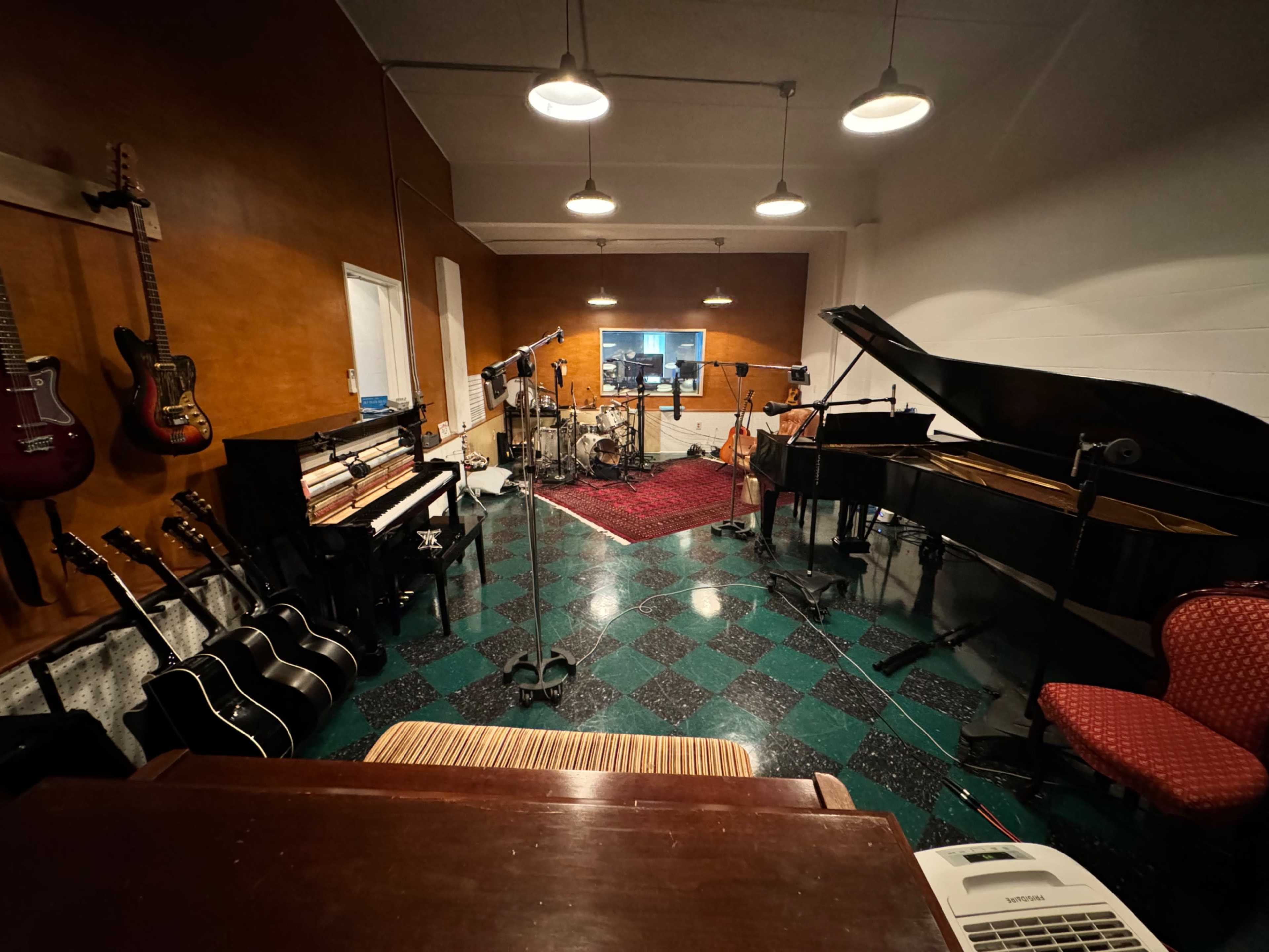 The image shows a music recording studio featuring various instruments, including guitars, a grand piano, and a drum set, arranged around a central area with sound equipment.