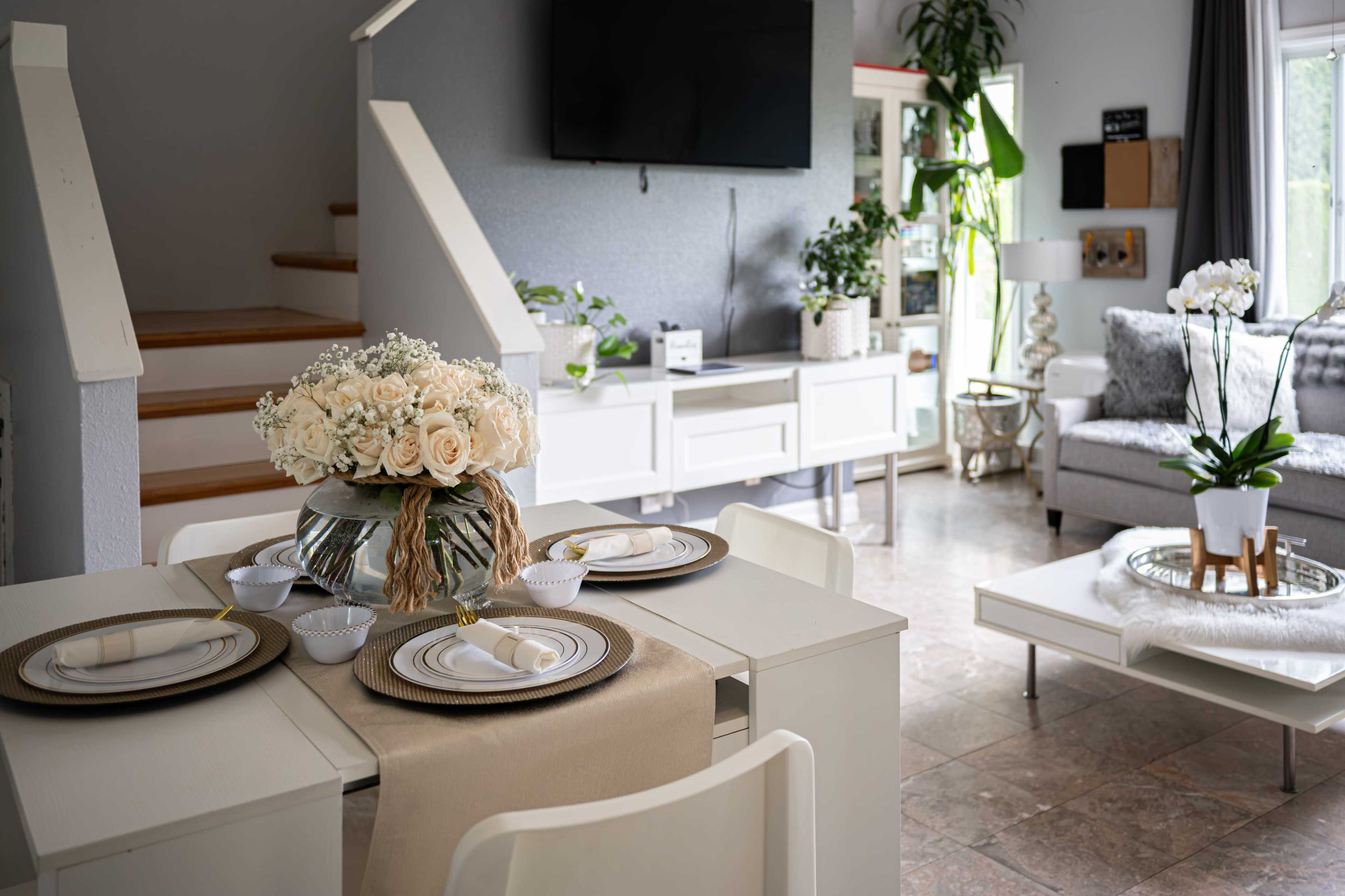 A dining area with a table set for a meal, featuring a floral centerpiece, near a living area with a sofa and plants.