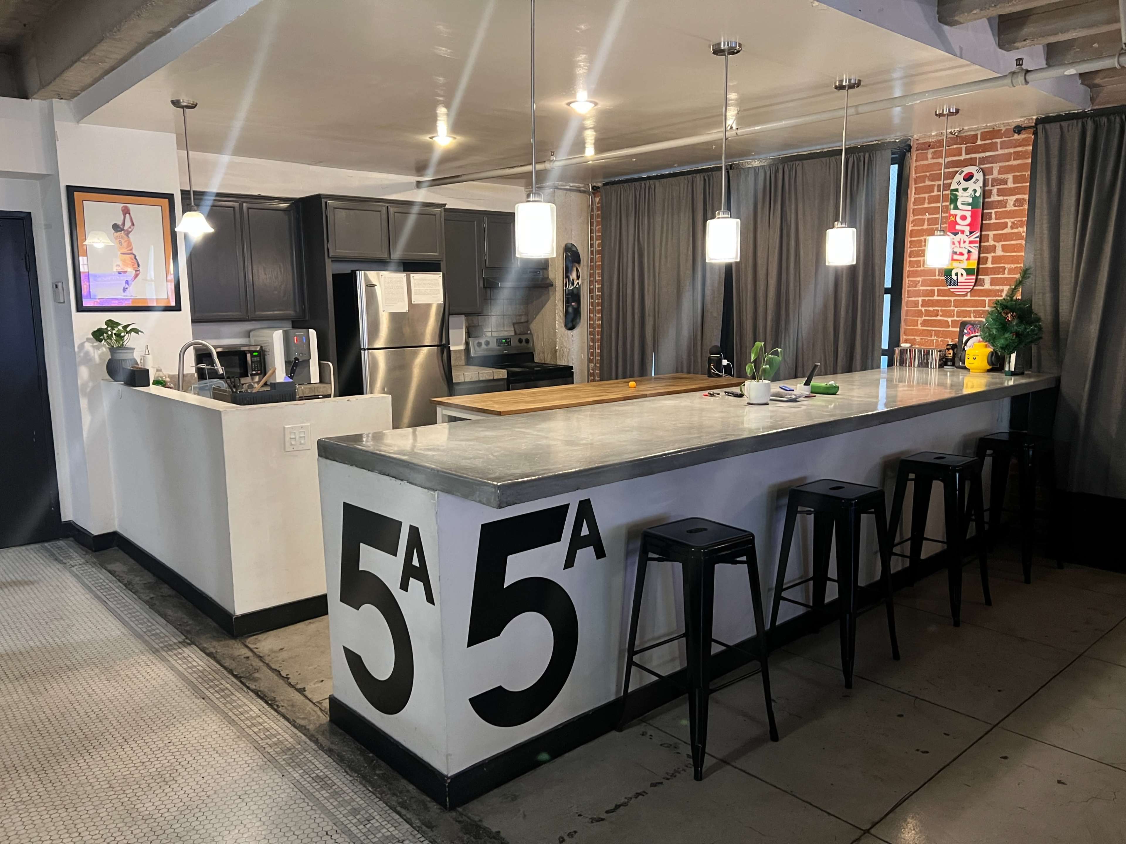 The image shows a modern kitchen with a concrete countertop, black cabinets, and pendant lighting, featuring a bar area with tall stools and a decorative plant.