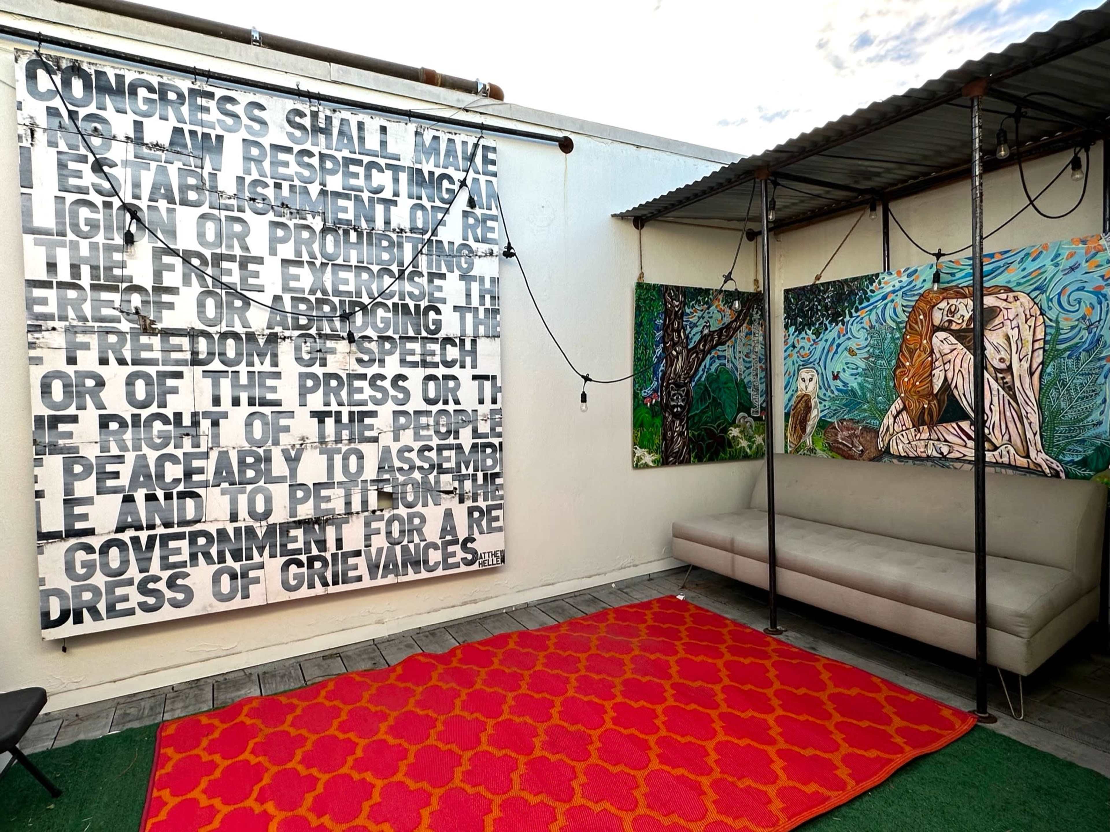 The image shows an outdoor space with a large artwork featuring text on one wall and colorful paintings on another, accompanied by a cozy seating area and a red patterned rug on the floor.