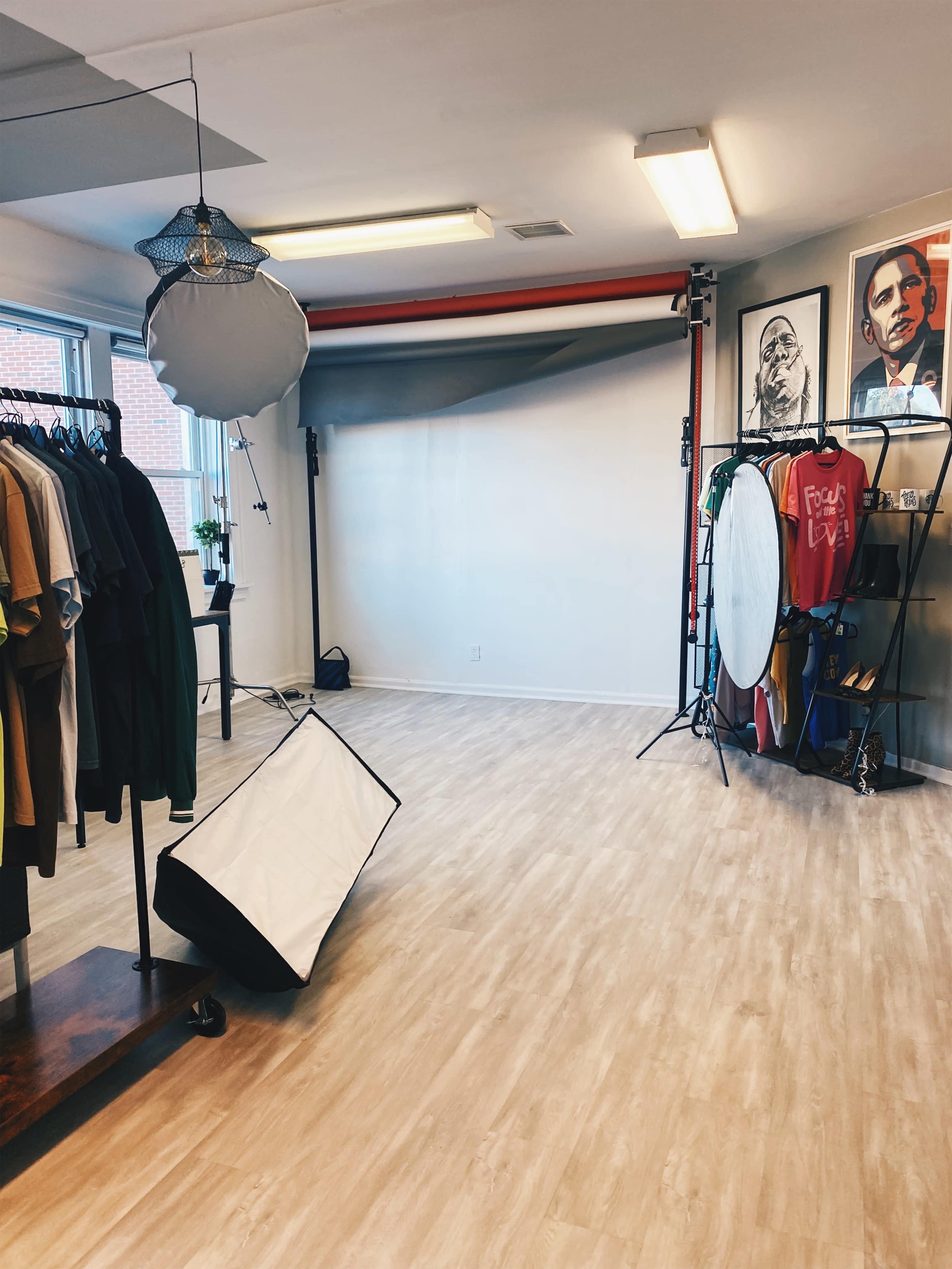 A photography studio featuring a backdrop, various lighting equipment, and clothing racks against light-colored walls.