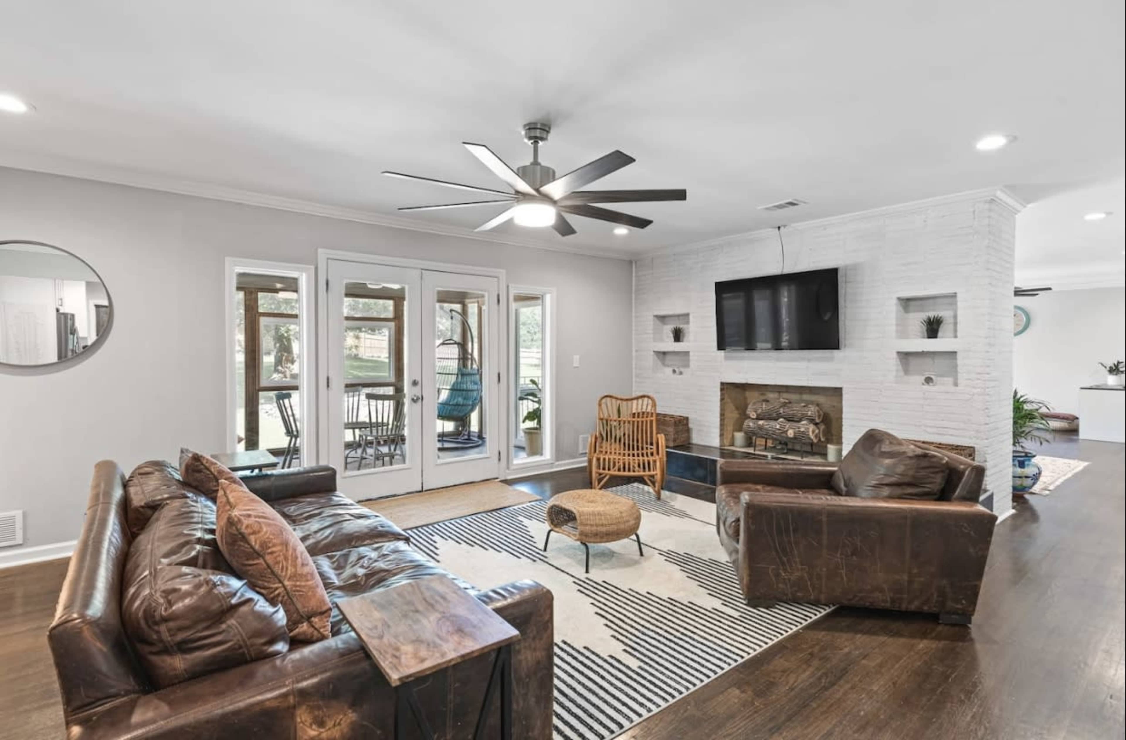 A modern living room featuring a brown leather sofa, a wooden coffee table, a stone fireplace, and large windows that provide natural light.