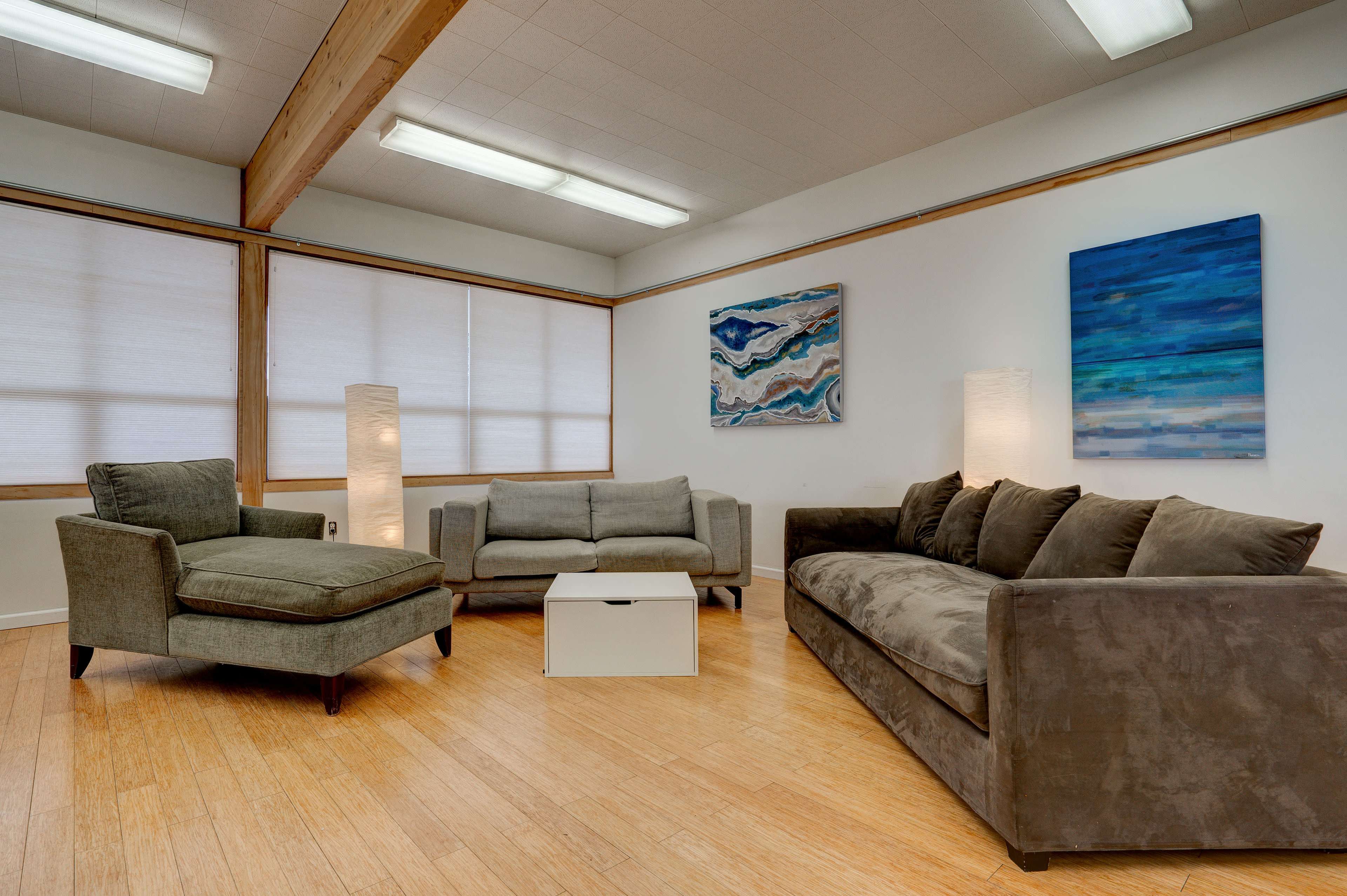 A modern living room with two sofas, an armchair, a coffee table, and wall art, illuminated by light fixtures and natural light filtering through the blinds.