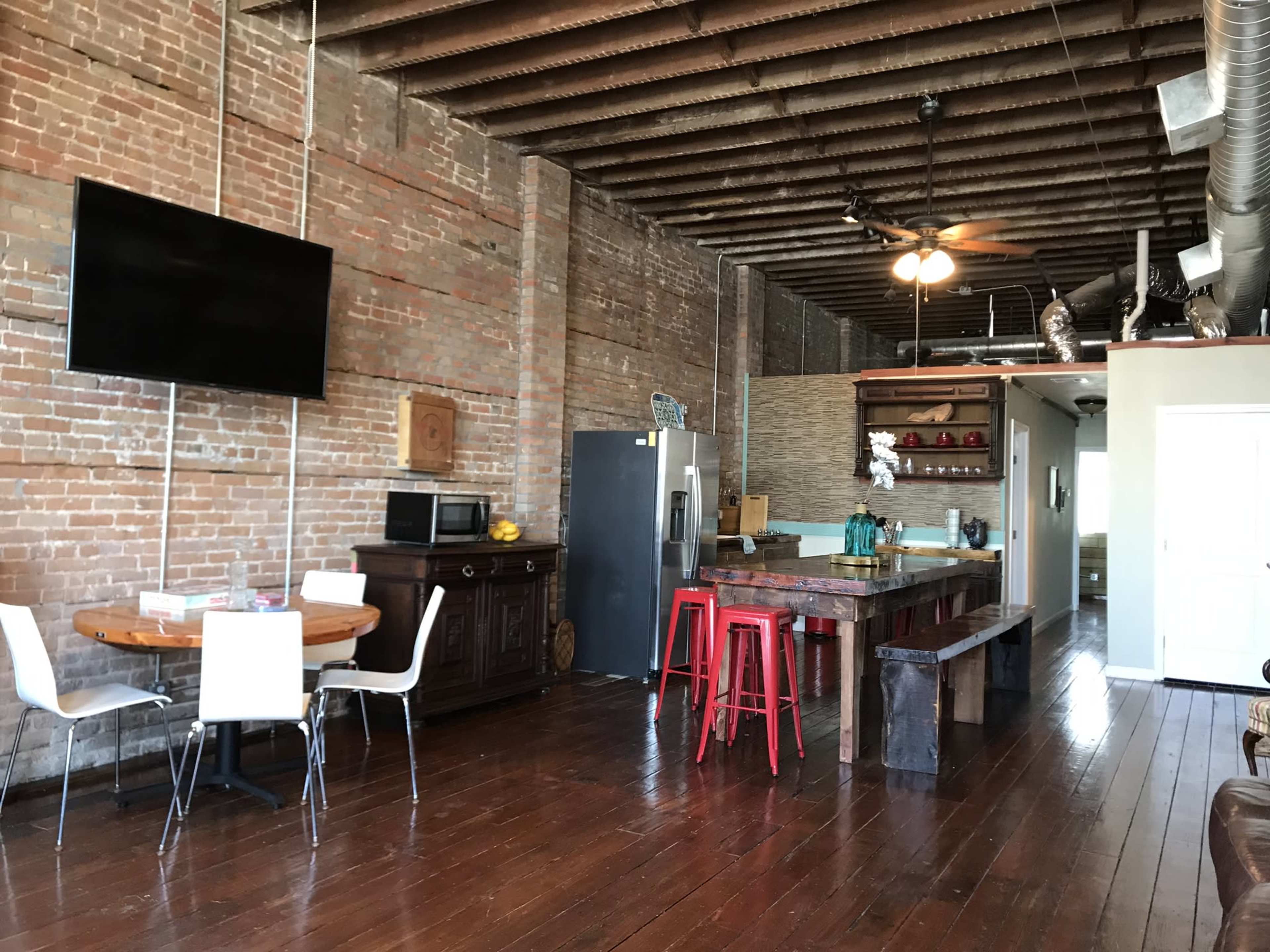 The image shows an industrial-style kitchen and dining area with exposed brick walls, wooden floors, and a mix of modern and rustic furnishings.