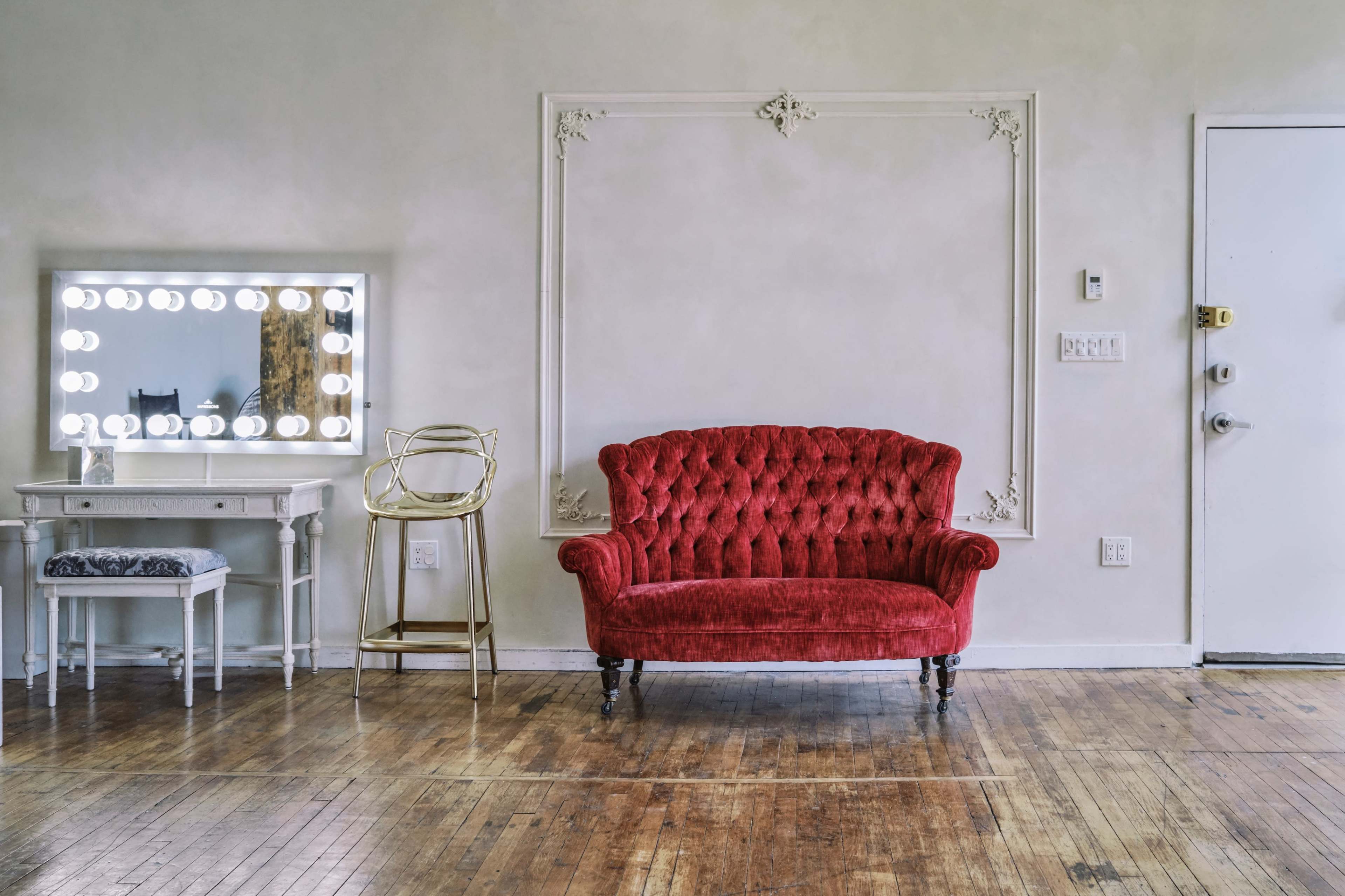 The image features a red tufted sofa positioned against a plain wall, with a mirrored vanity and a gold-accented chair nearby, all set on a wooden floor.