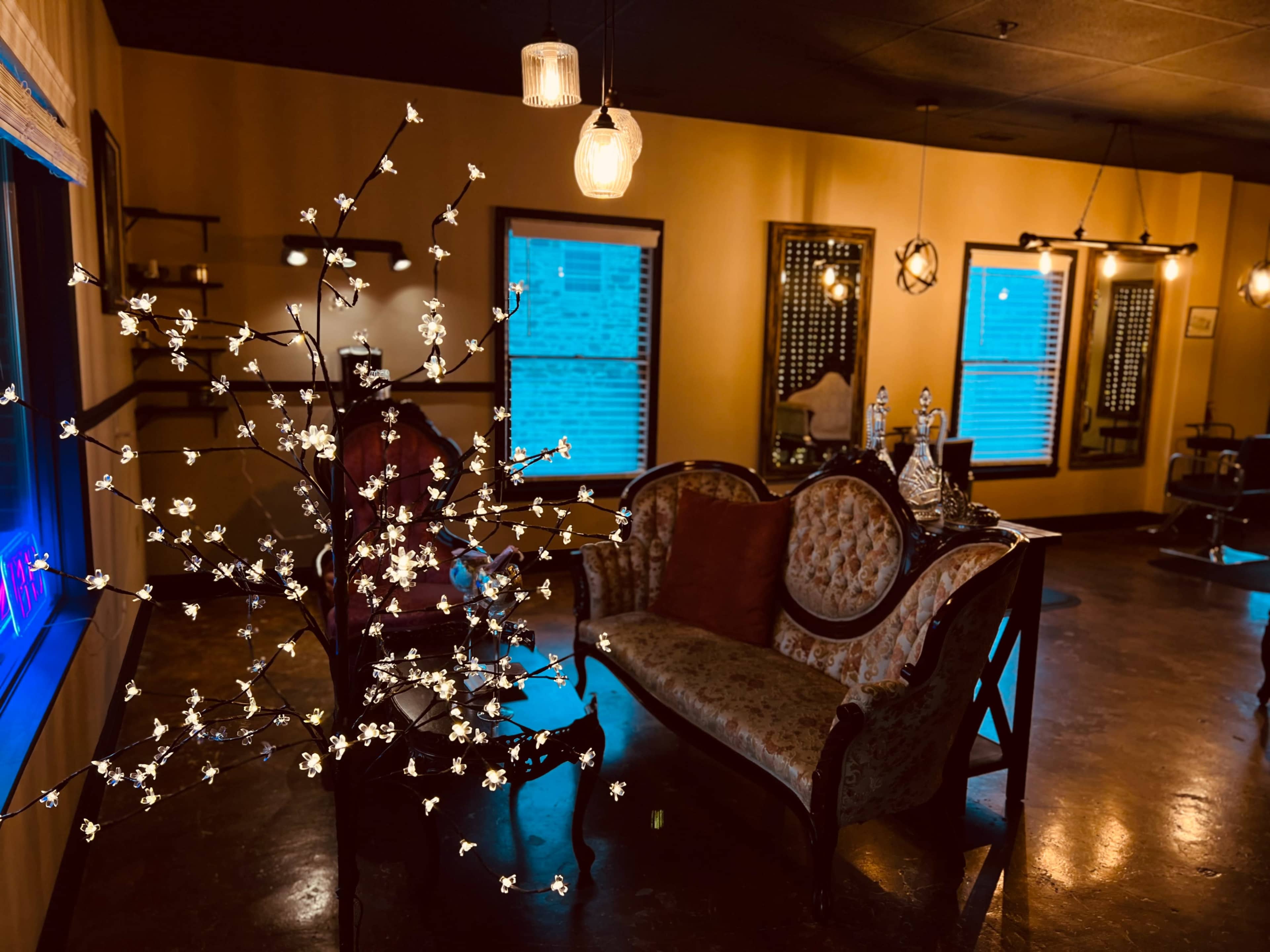 The image shows a dimly lit salon with vintage furniture, including a patterned sofa and a decorative tree with small flowers, alongside illuminated windows.