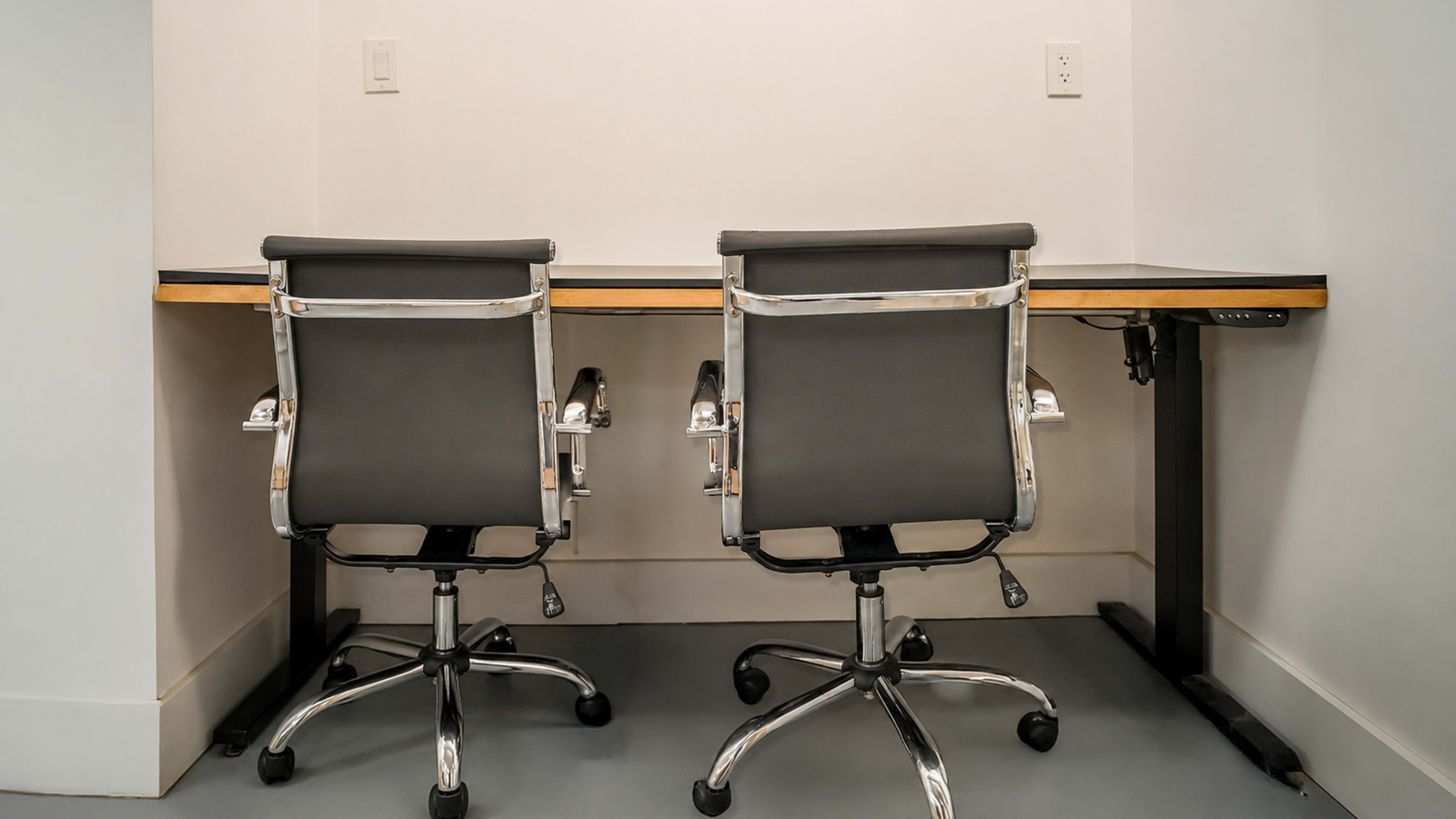 The image shows a minimalist office space with two black ergonomic chairs positioned in front of a wooden desk against a light-colored wall.