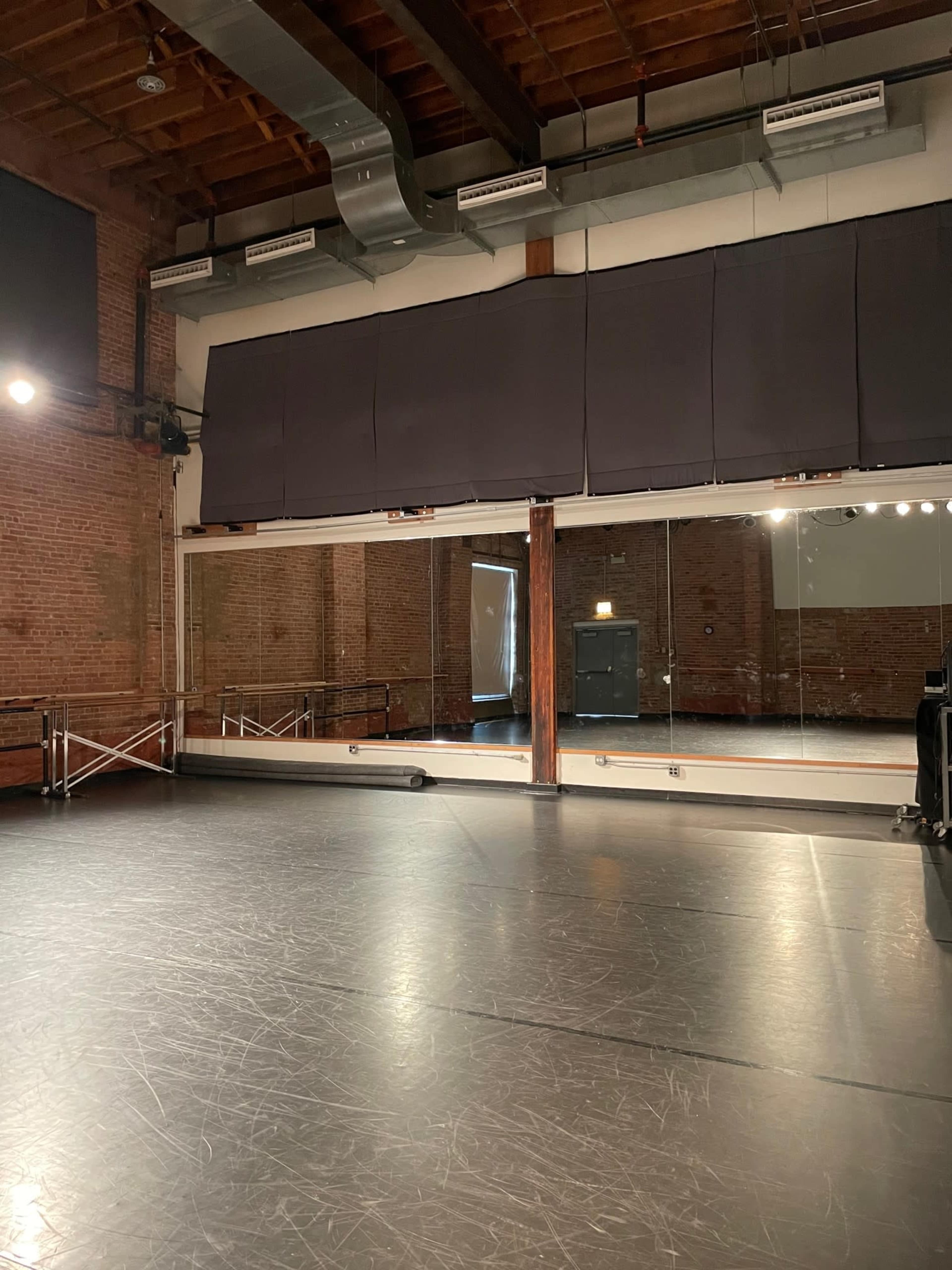 A dance studio with a wooden ceiling, brick walls, a mirrored wall, and a dark flooring surface.