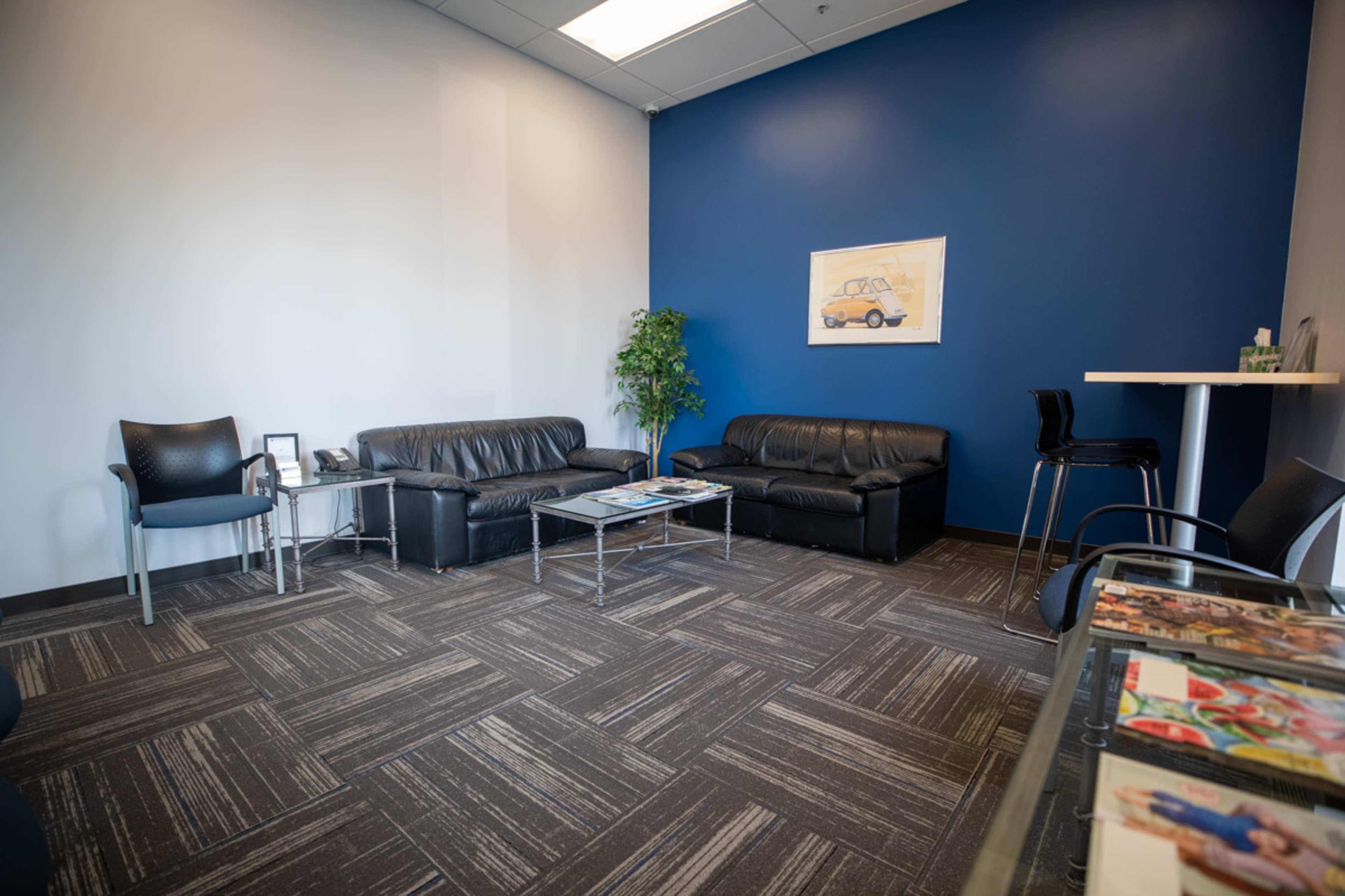 A modern waiting room with black leather sofas, a small coffee table, a tall barstool table, and a decorative plant against a blue and white wall.