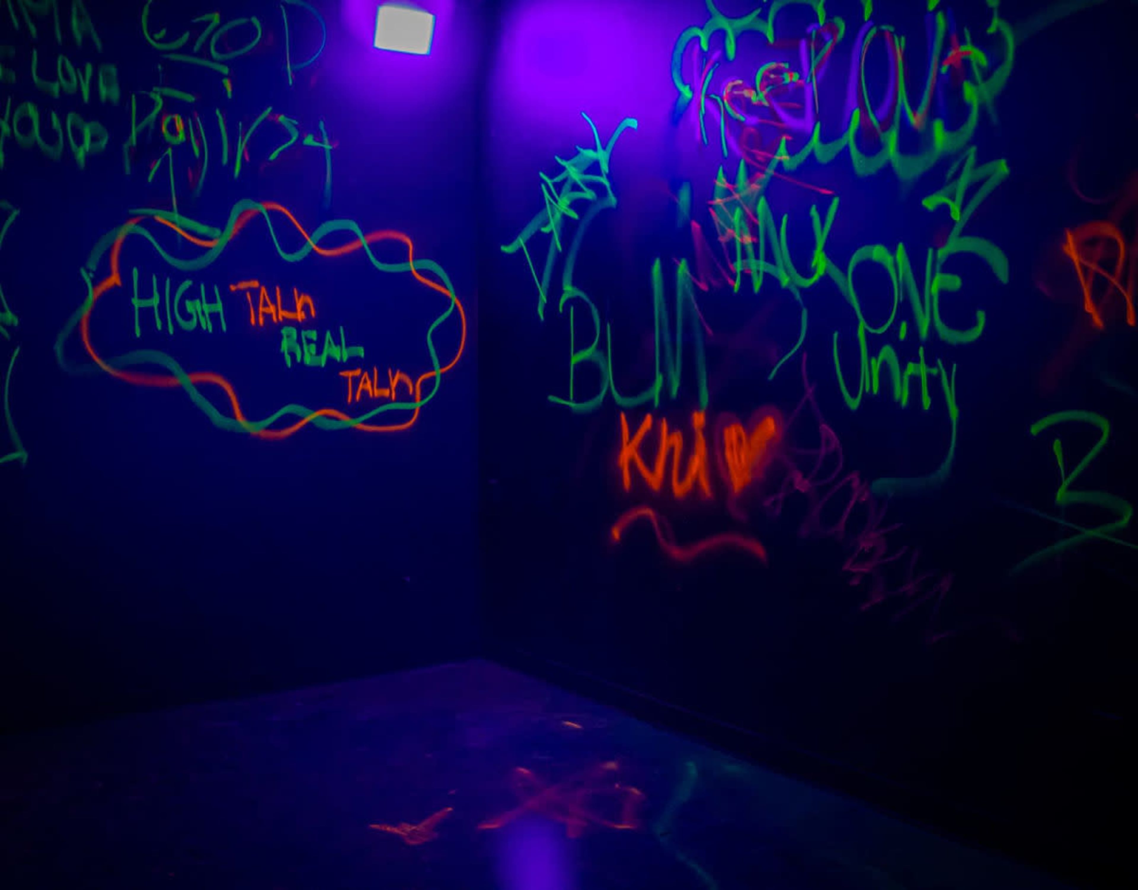 The image shows a dark room with colorful graffiti on the walls, including phrases like "HIGH TALK REAL TALK" and "UNITY."