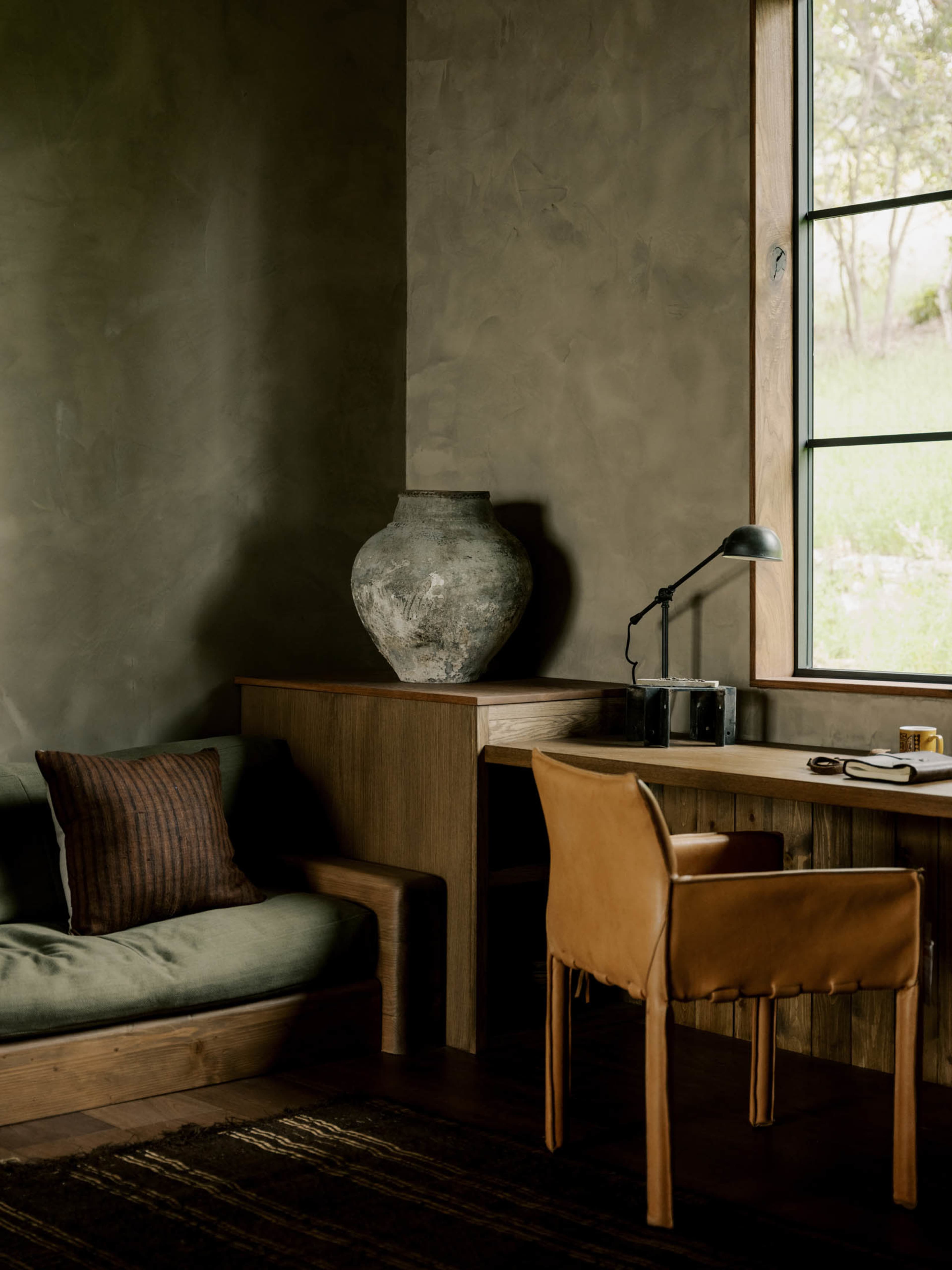 The image shows a cozy room with a wooden desk and chair, a green sofa, an artistic vase, and a large window that lets in natural light.