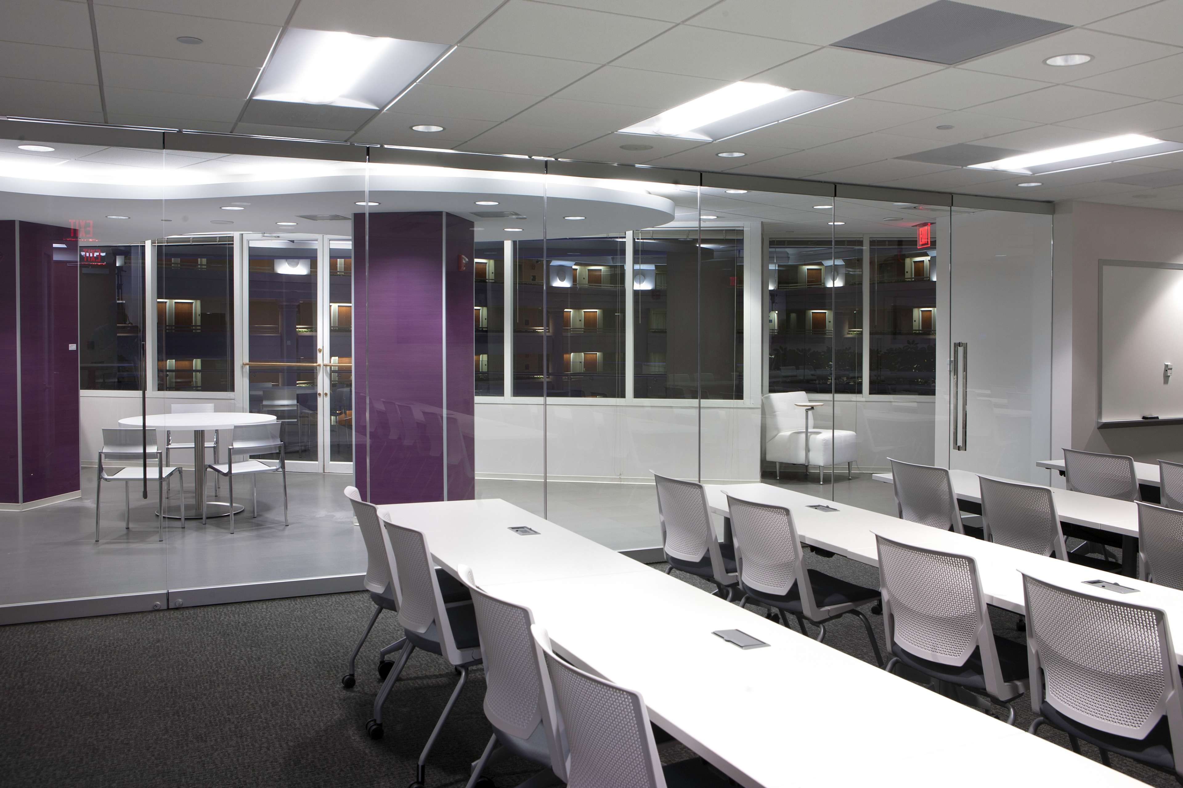 A modern office space with white tables and chairs arranged in a classroom style facing a glass-walled conference area and large windows overlooking a cityscape.