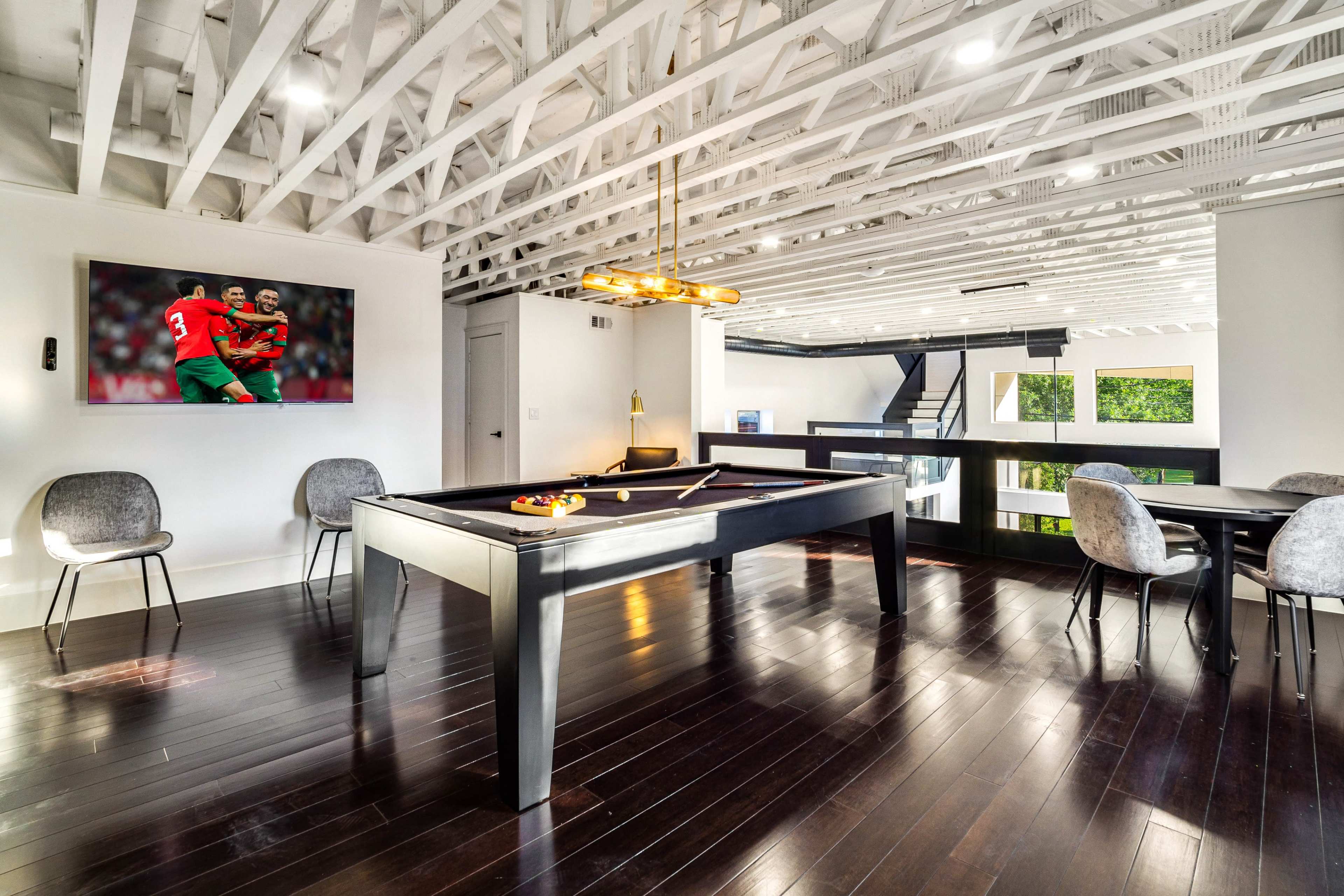 The room features a pool table, seating area, and a mounted television displaying a soccer match, all beneath a wooden beam ceiling.