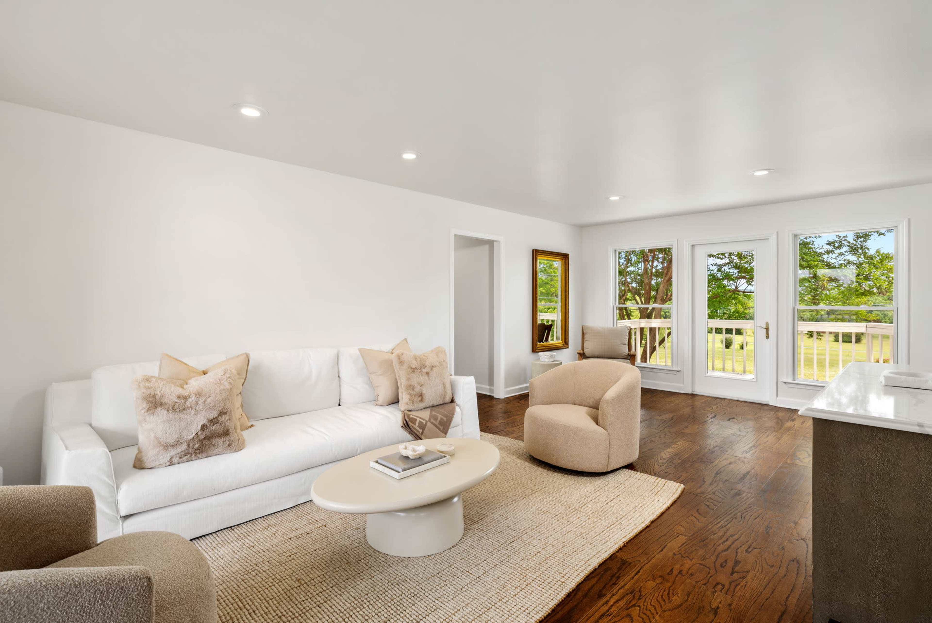 The image shows a bright living room with a white sofa, two light-colored chairs, a round coffee table, and large windows that open to a view of greenery outside.