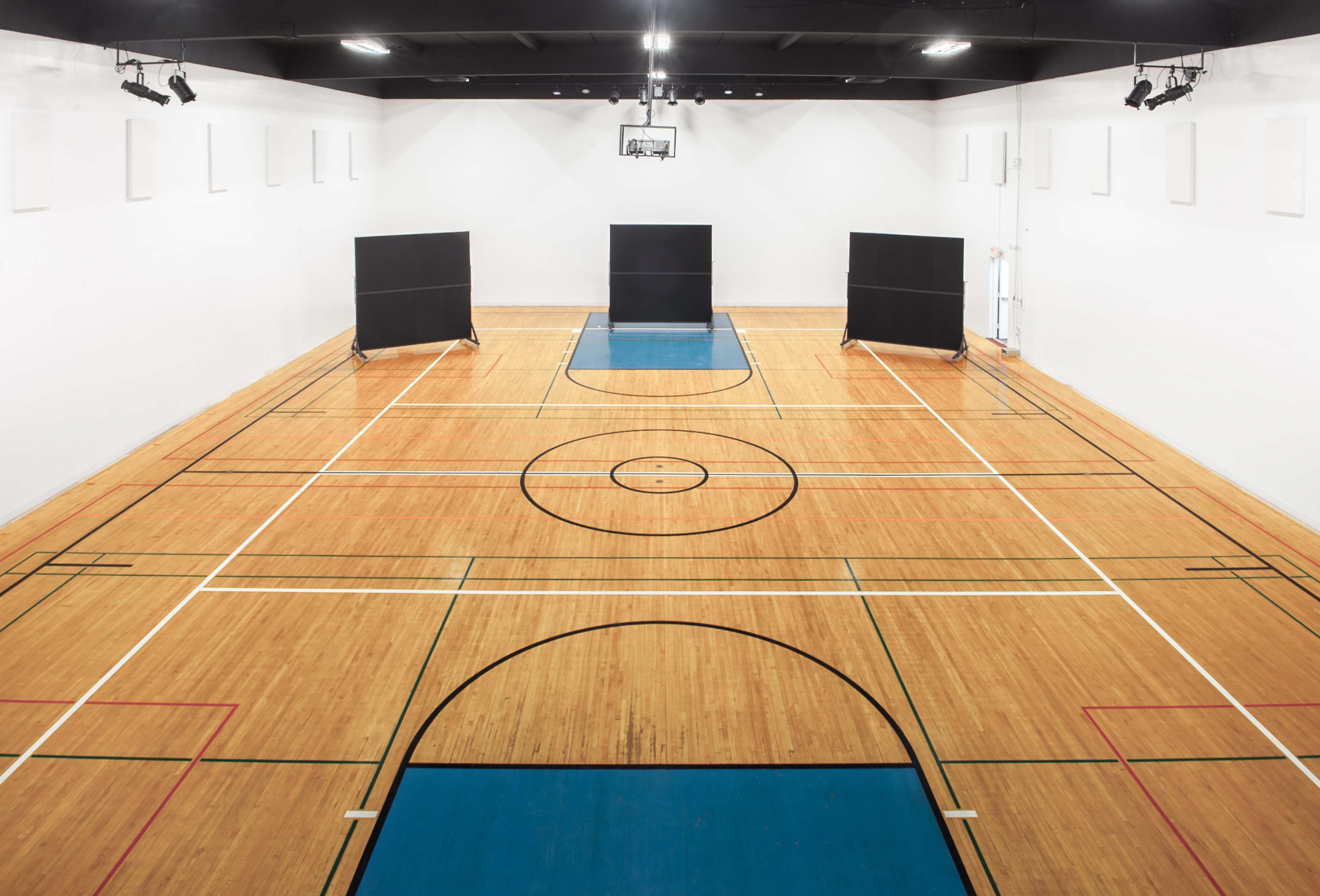 The image shows a large, empty indoor basketball court with a hardwood floor, marked lines, and two black dividers at each end.