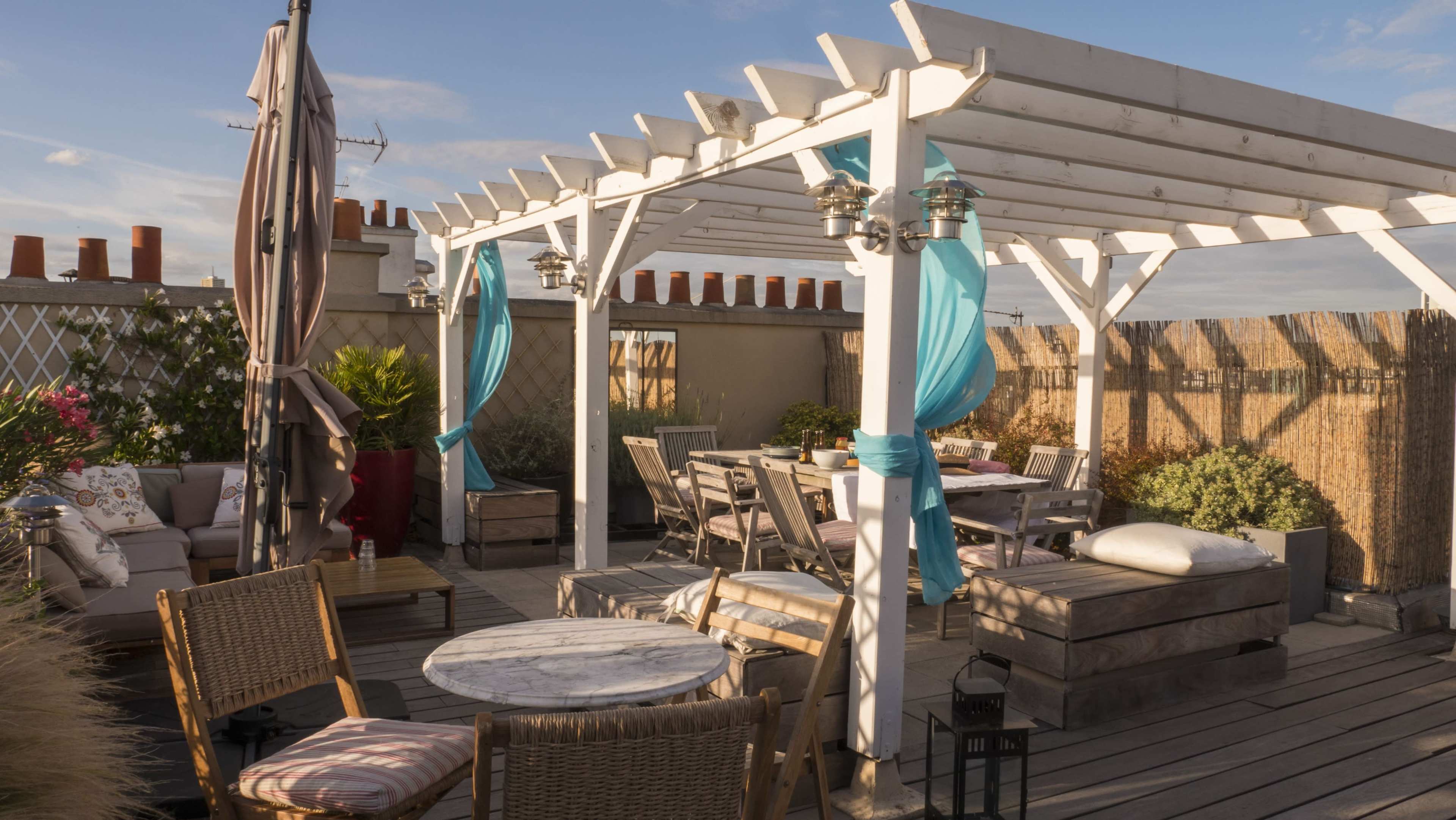 A rooftop patio features a white pergola adorned with blue fabric drapes, surrounded by wooden furniture and decorative plants.