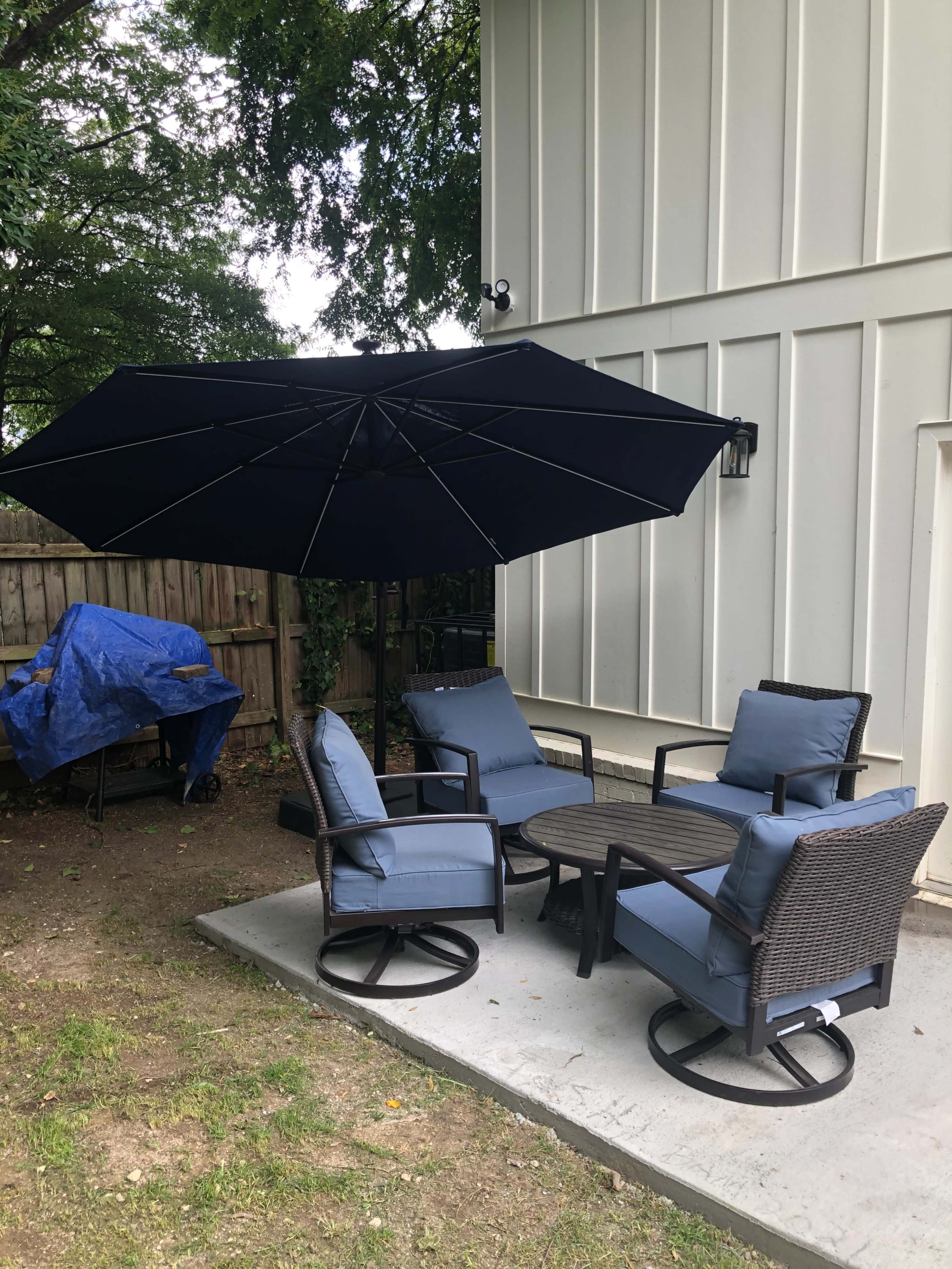 A patio area features four blue cushioned chairs around a circular table, shaded by a large black umbrella, with a grill partially covered by a blue tarp nearby.