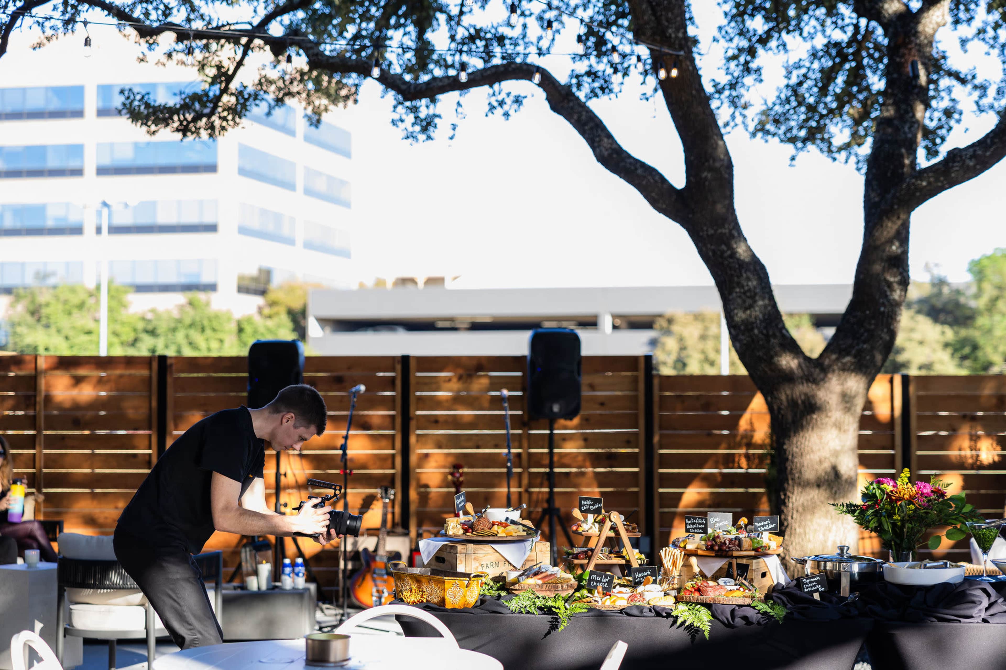 A person is taking photographs of a stylish outdoor food display set up under a large tree, with a modern building visible in the background.
